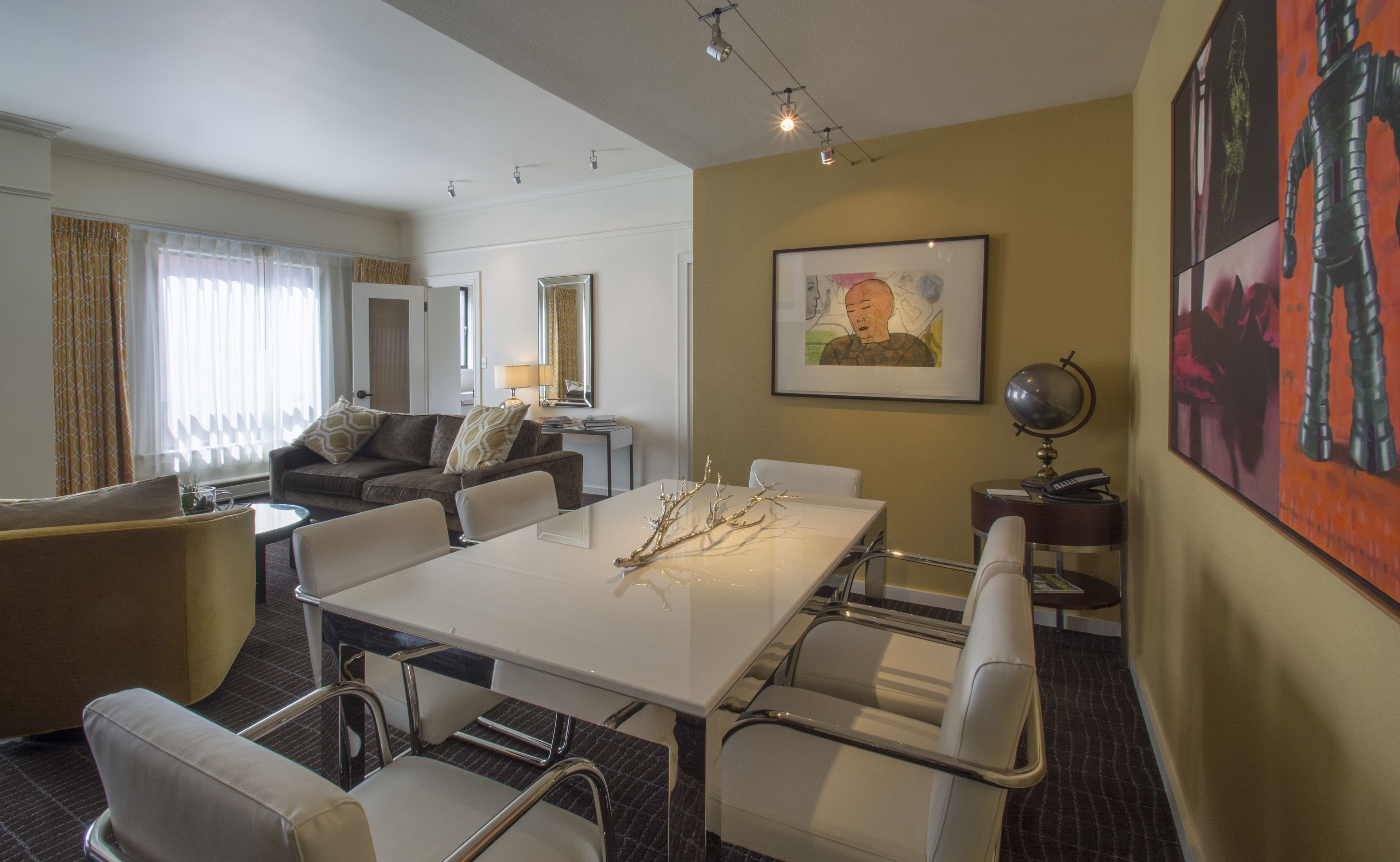 A hotel room with a dining table, chairs, a sofa, a lamp, and a painting on the wall.