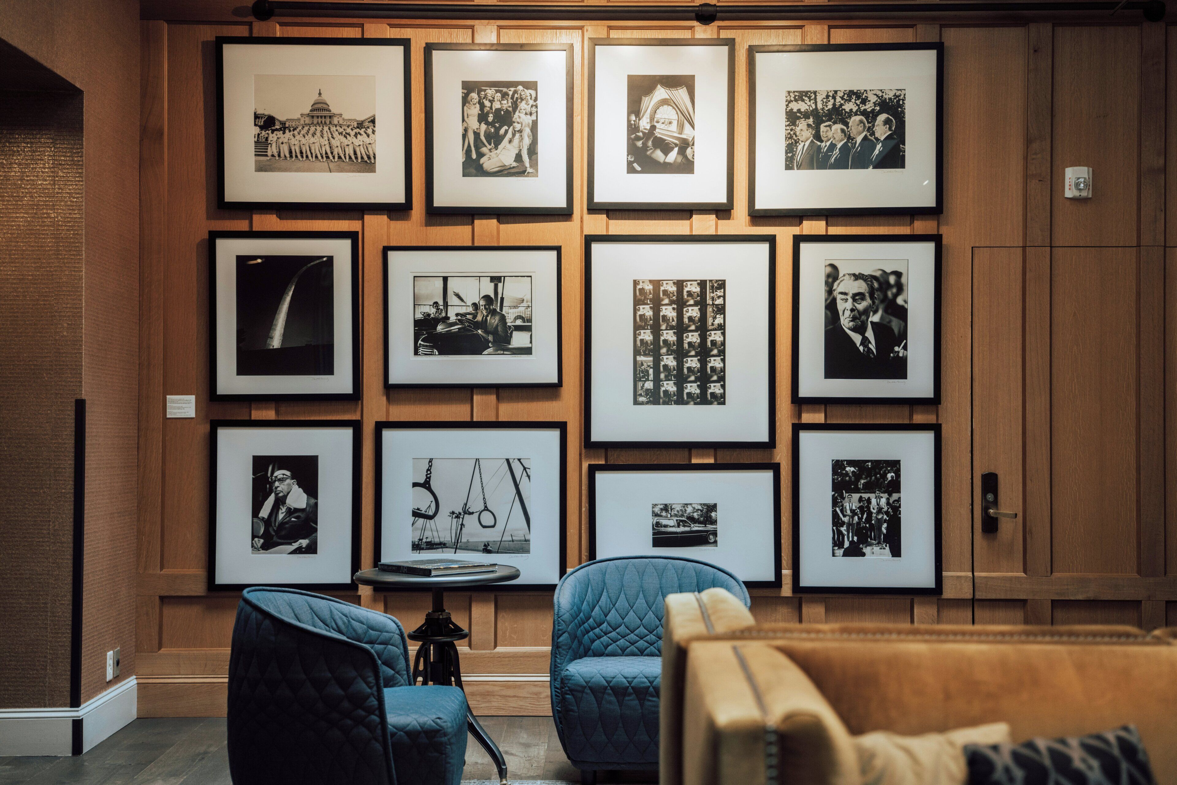 Una pared con fotografías blanco y negro enmarcadas, una mesa redonda con un libro y dos sillas azules.