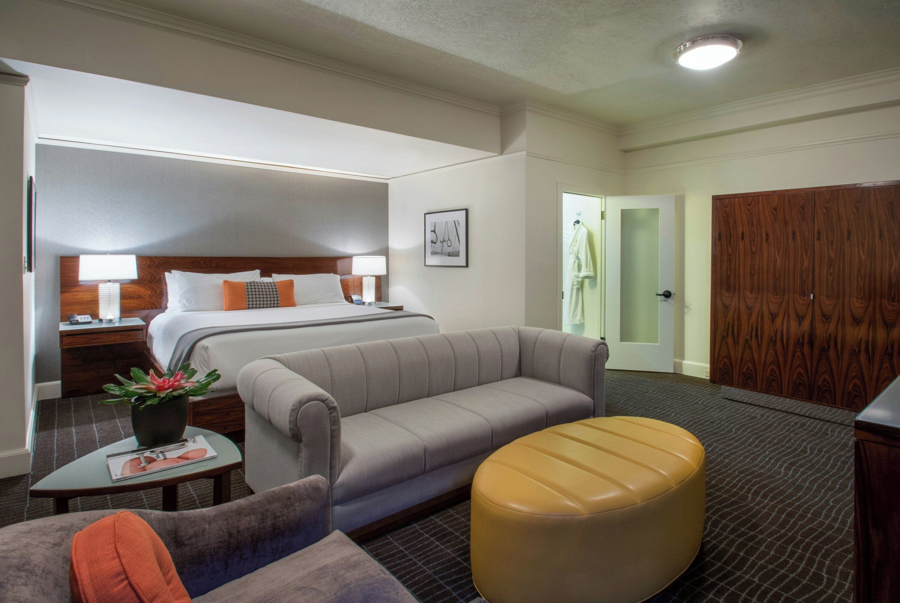 A hotel room with a bed, a sofa, an ottoman, a small table with a plant, and a nightstand with a lamp.