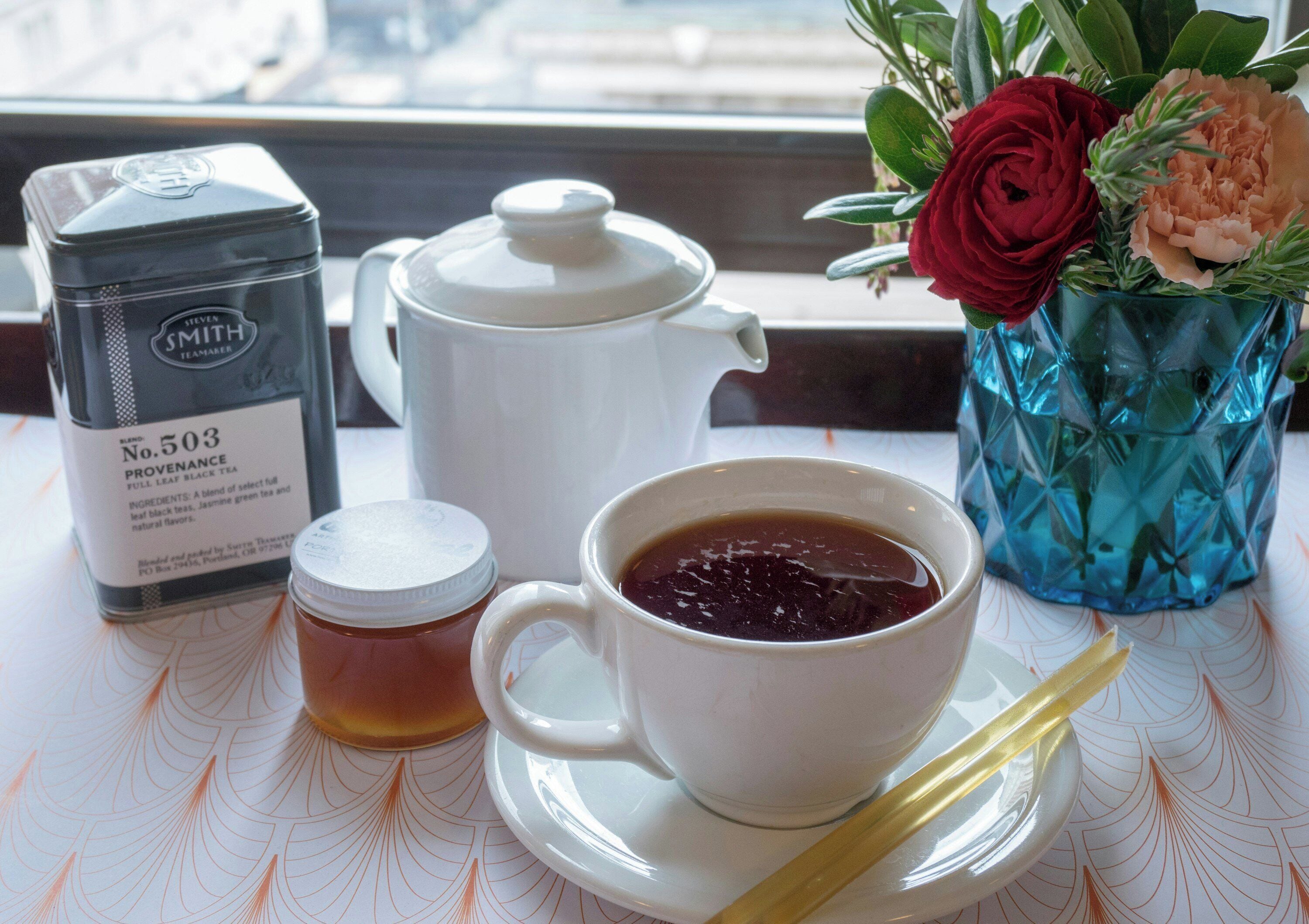 A tea set with a teapot, a cup, and a saucer, a jar of honey, and a box of tea on a table with a window view.