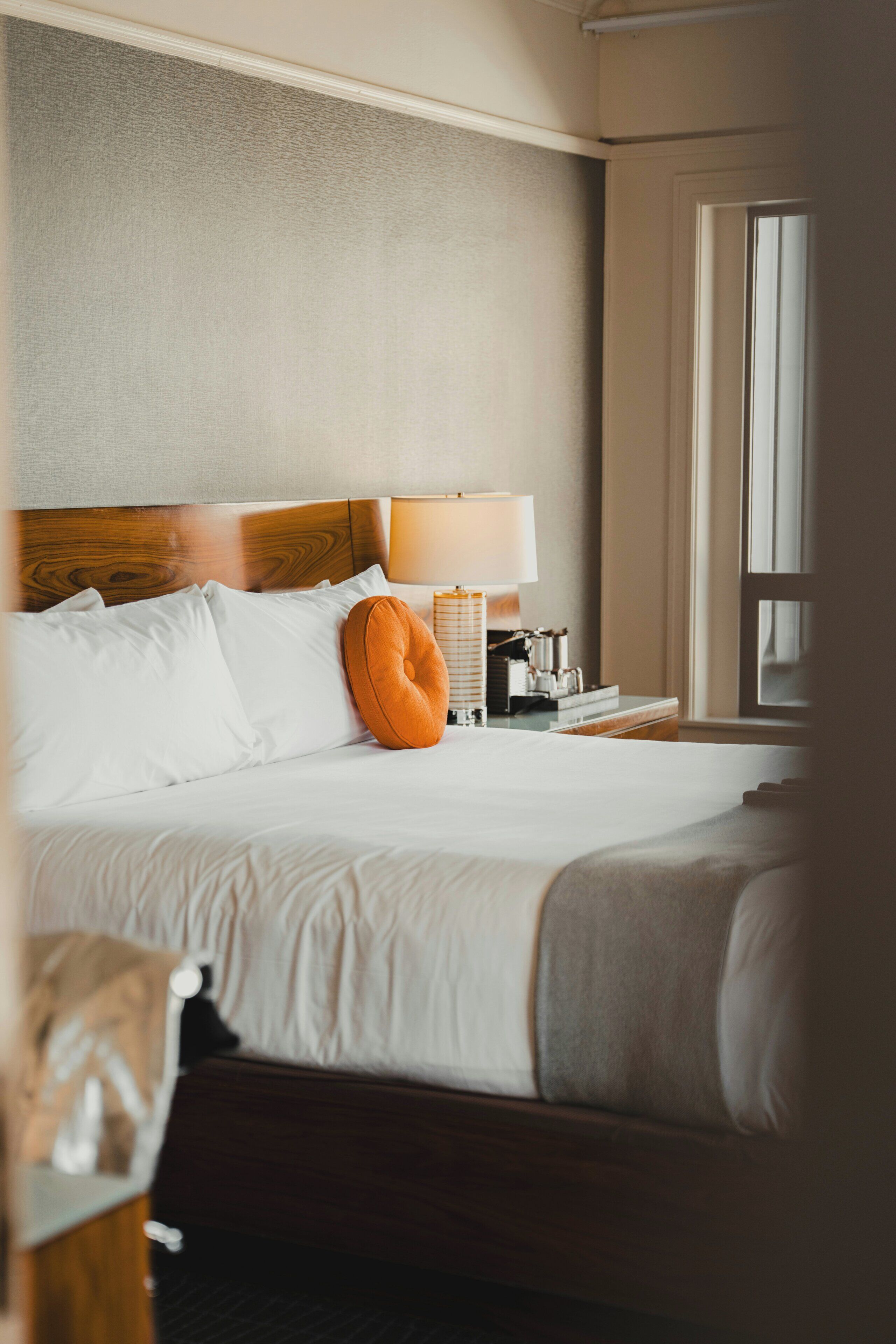 Una habitación de hotel con cama, almohadas, lámpara, mesita de noche y ventana.