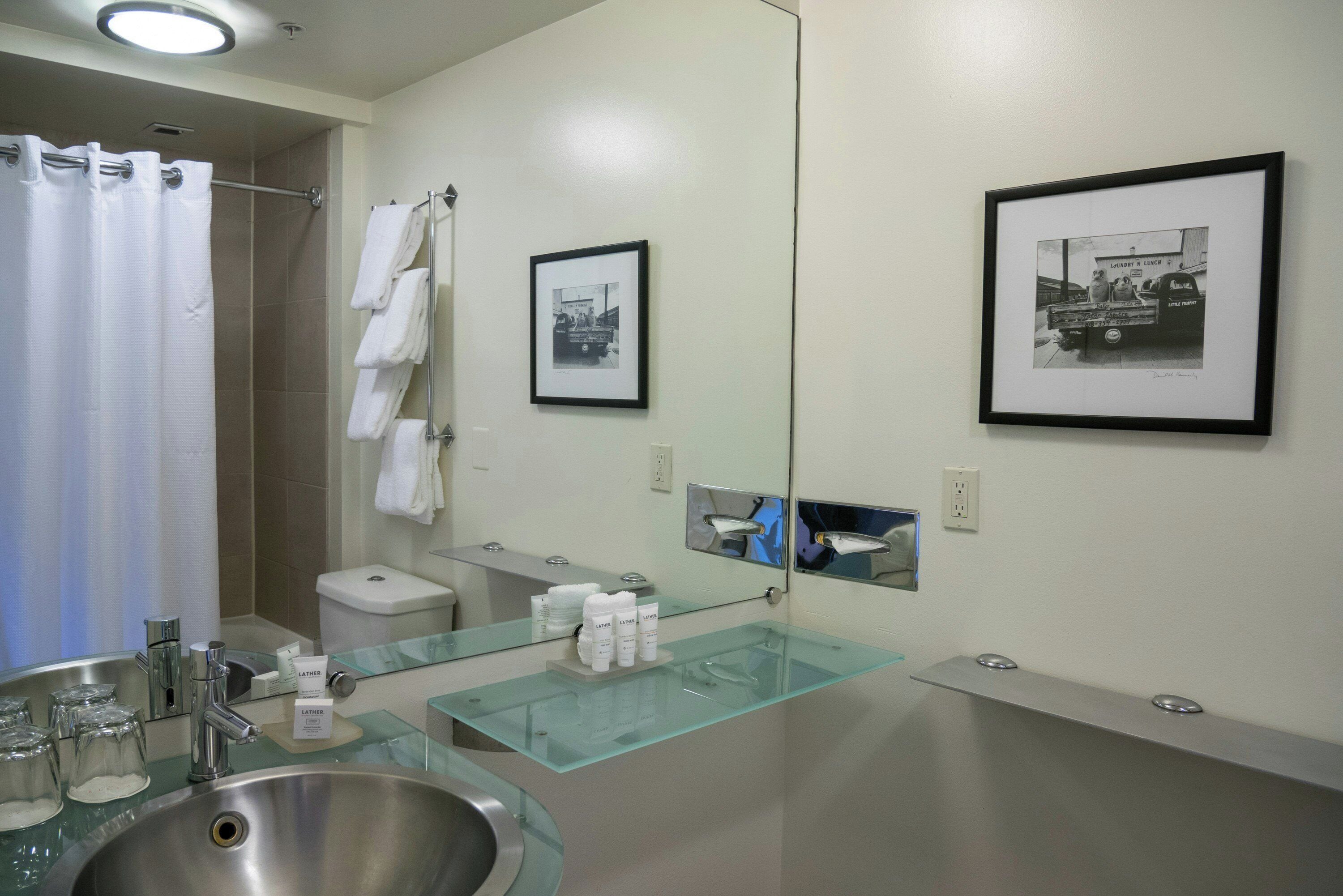 A modern bathroom with a glass sink, a shower, and two framed photographs on the wall.
