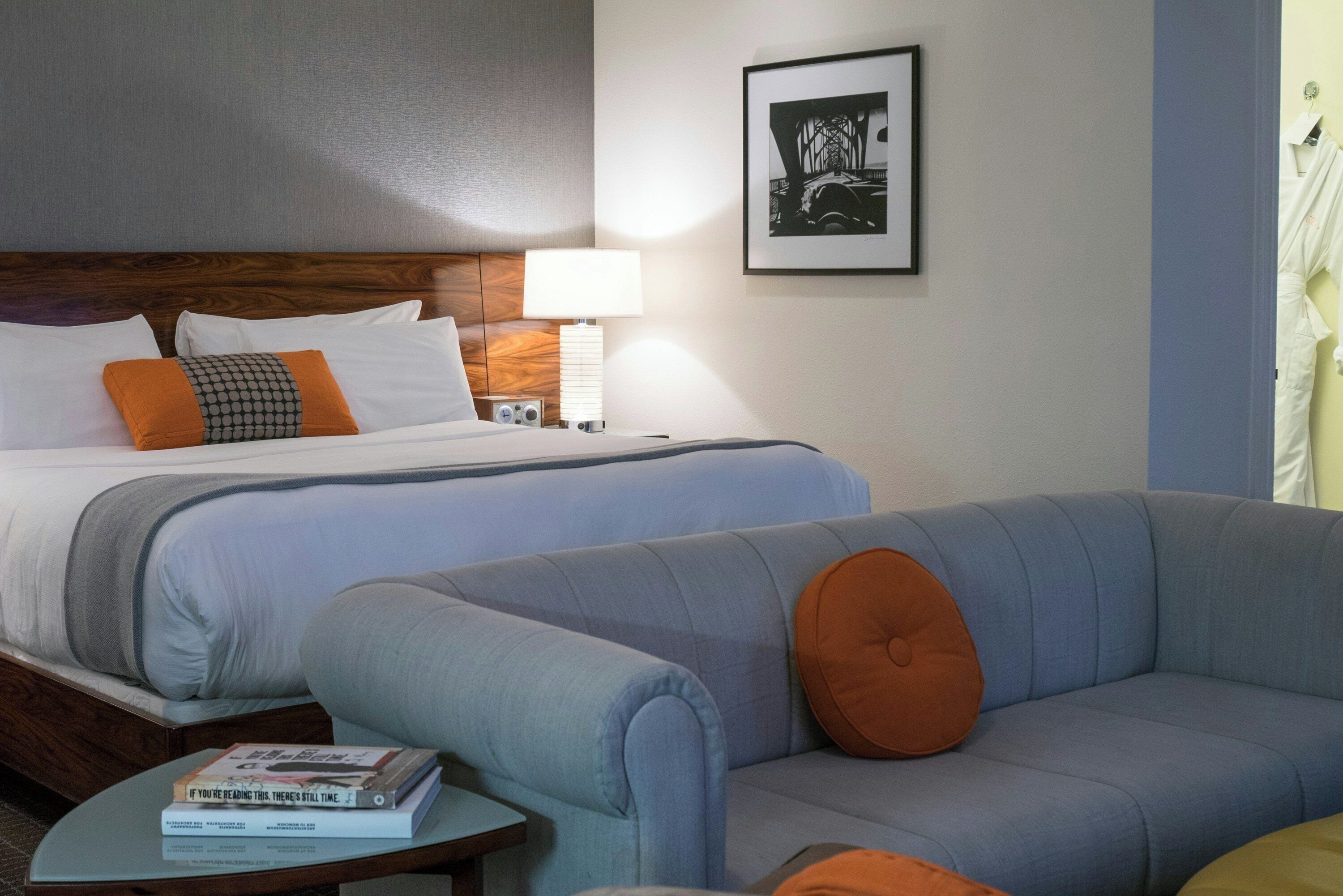 A hotel room with a bed, two pillows, a bedside table with a lamp, a framed picture on the wall, and a sofa with a cushion.