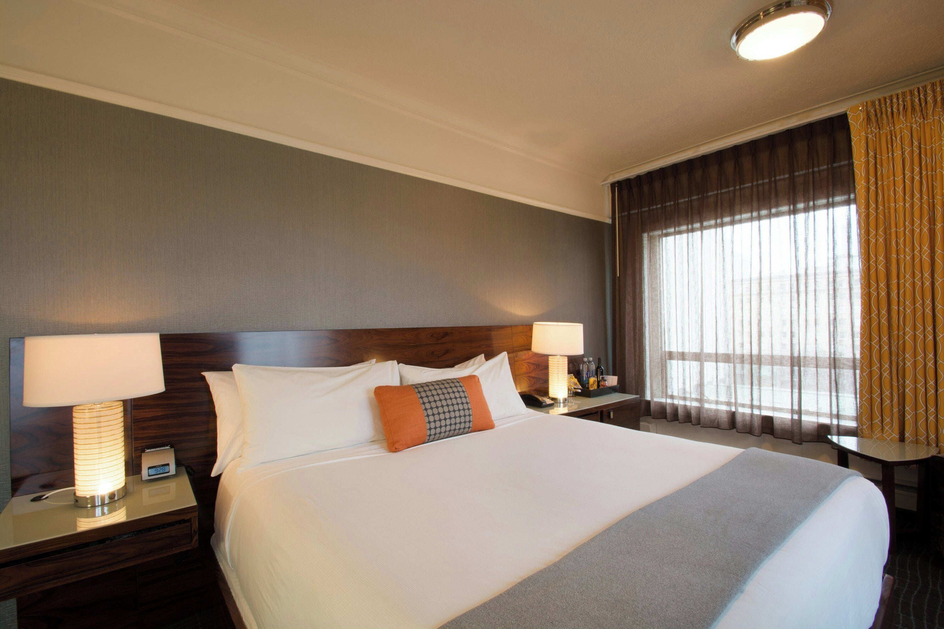 A hotel room with a large bed, two bedside lamps, a nightstand, a window with curtains, and a ceiling light.