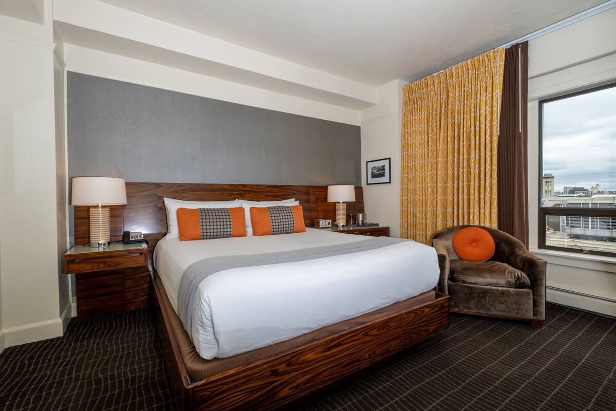 A hotel room with a large bed, two bedside tables, a chair, and a window with a city view.