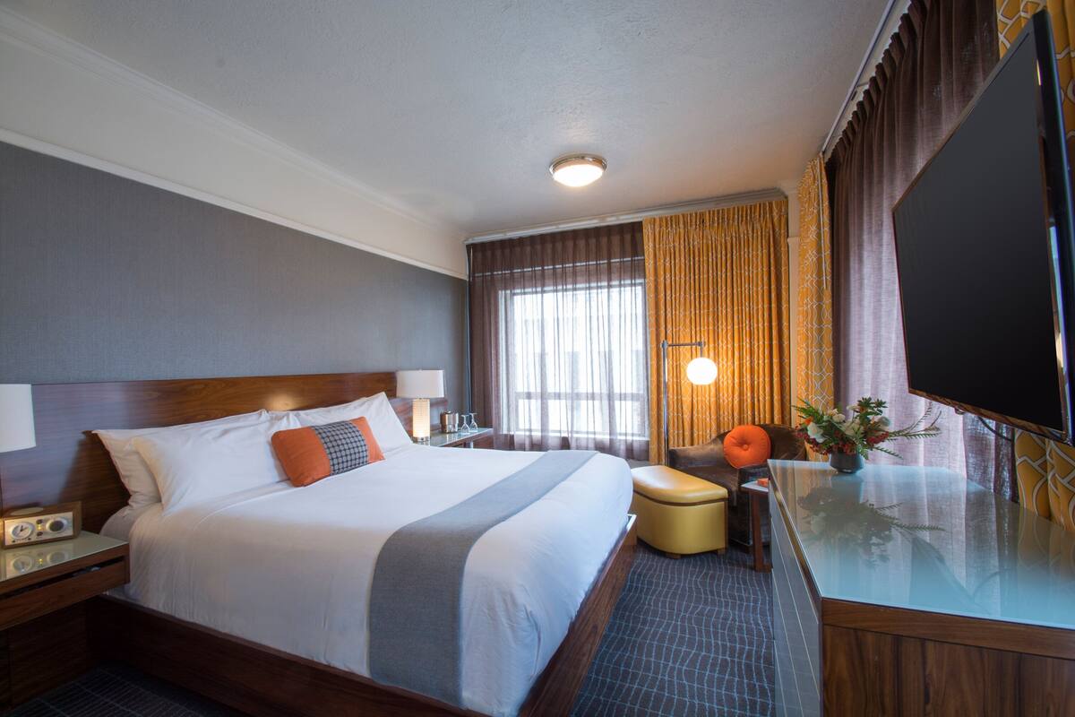 A hotel room with a large bed, a desk with a flat-screen TV, a chair, and a small table with a vase of flowers.