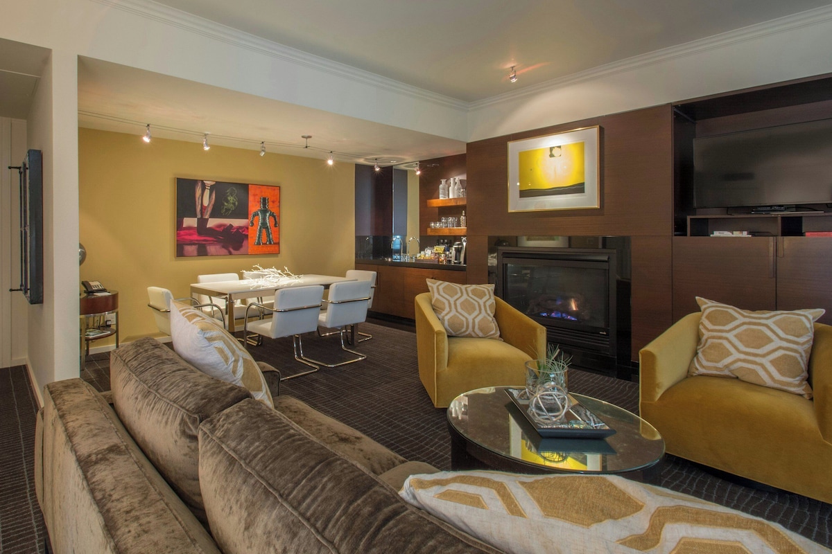 A living room with a sofa, armchairs, a coffee table, a dining area with chairs, a fireplace, and a television.
