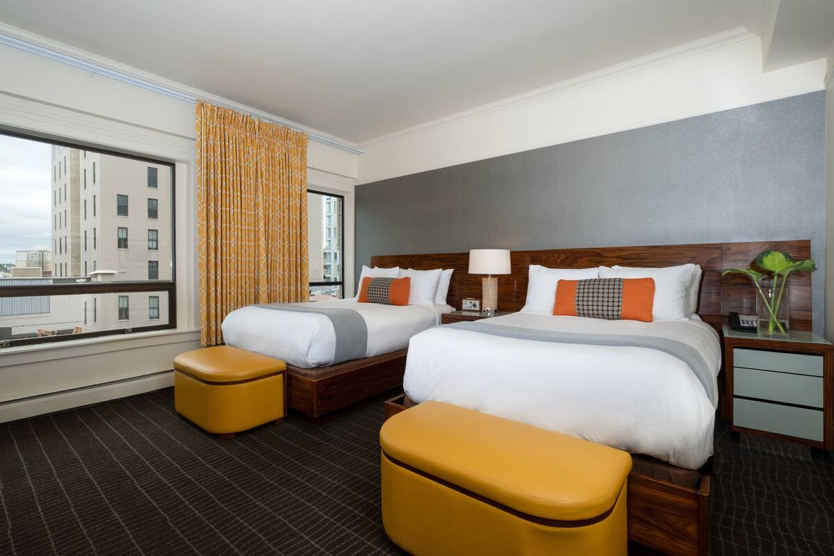 A hotel room with two beds, a large window with a view of buildings, and a cityscape.