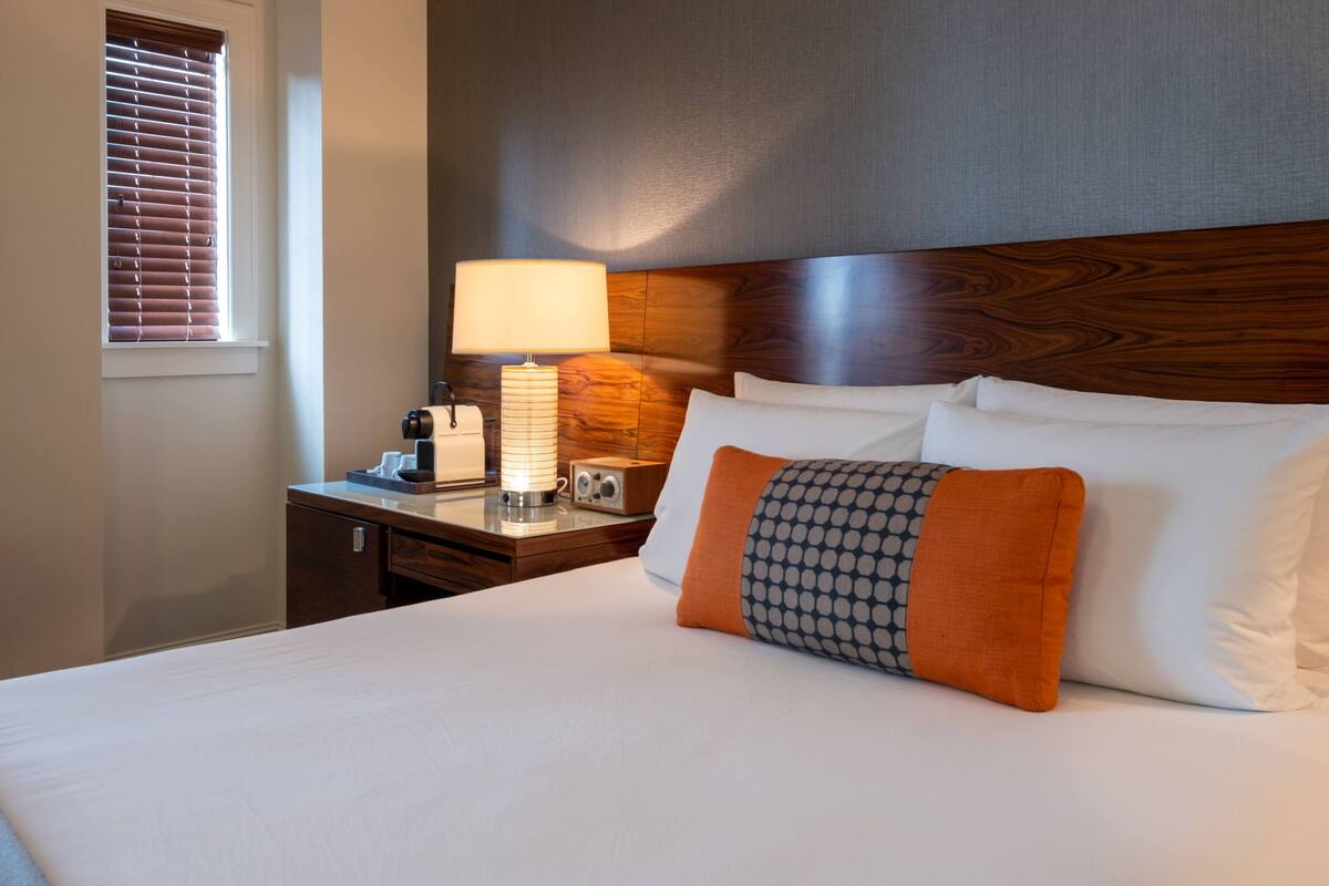 A hotel room with a neatly made bed, a bedside lamp, a radio, and a window with blinds.