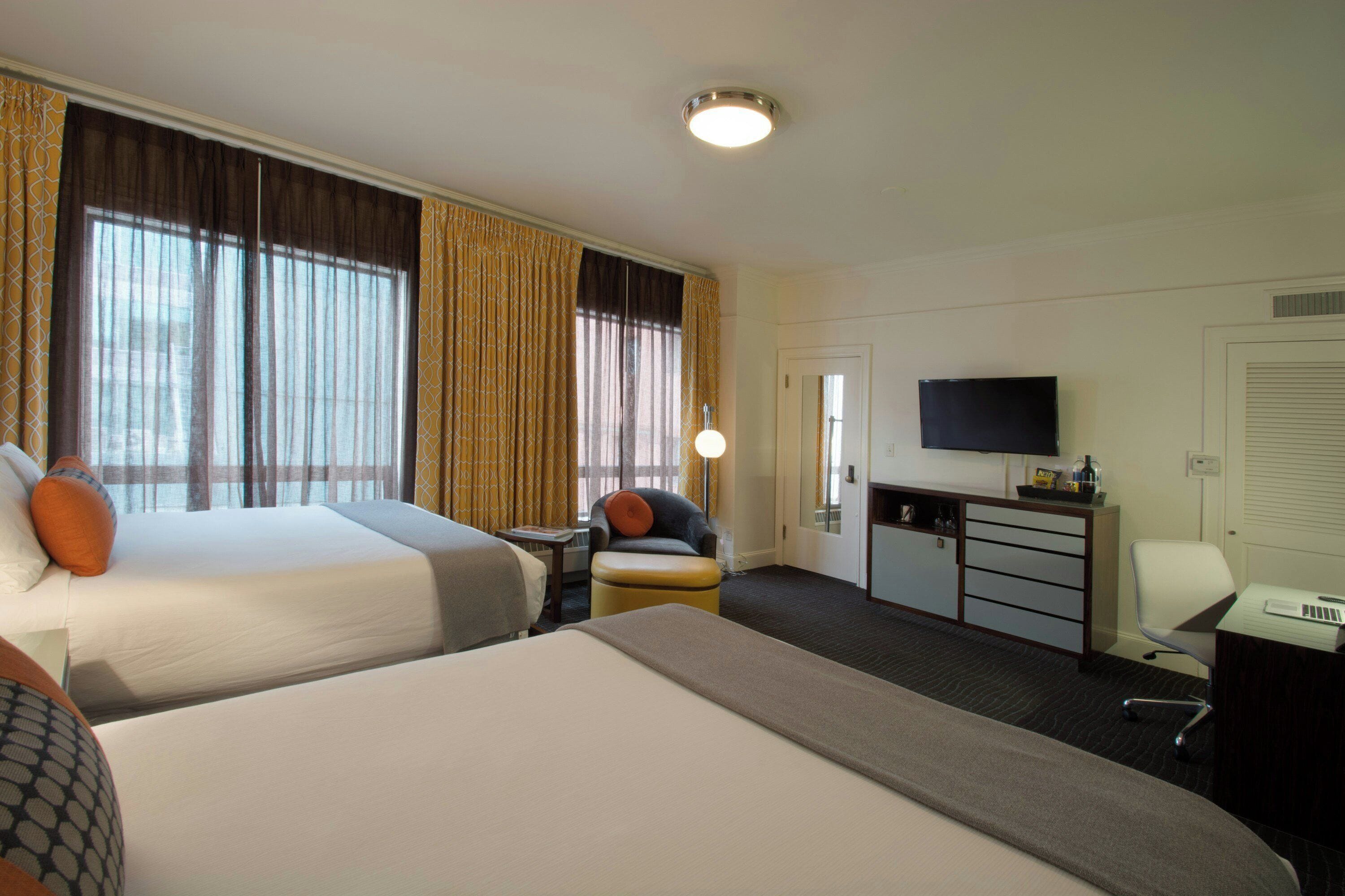 A hotel room with two beds, a desk, a chair, a television, and a minibar.