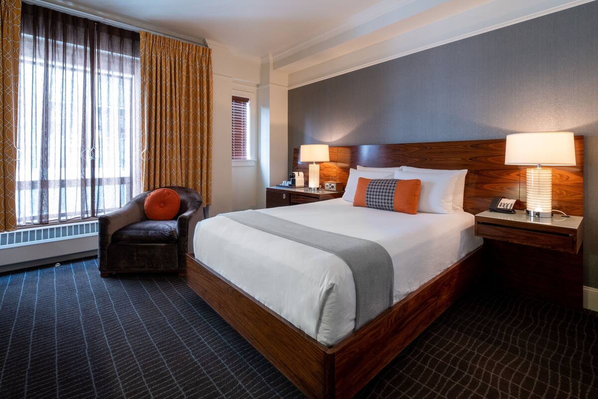A hotel room with a large bed, two bedside tables with lamps, a chair with an orange cushion, a window with curtains, and a radiator.