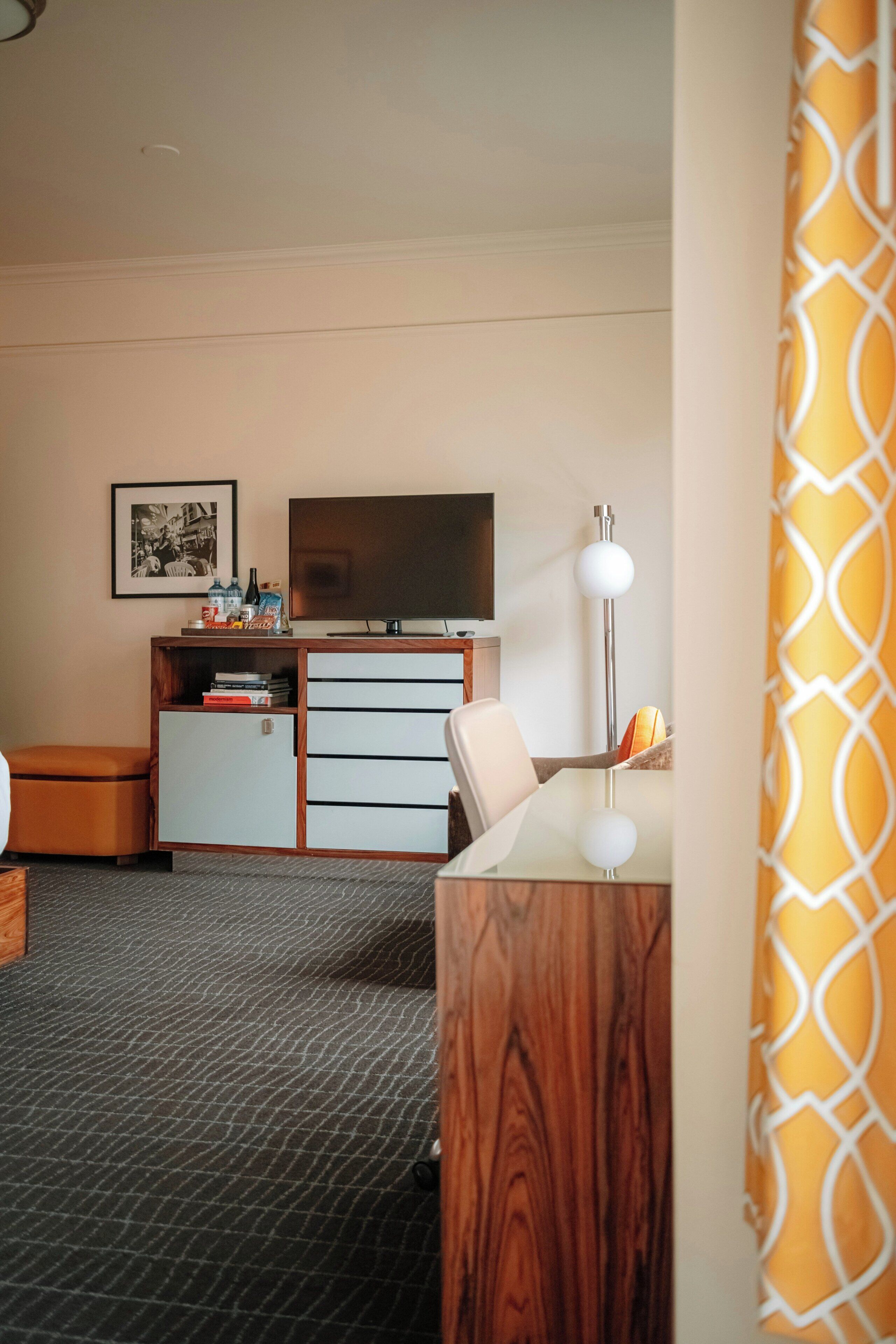 A hotel room with a television, a desk, a chair, a small cabinet, and a patterned carpet.