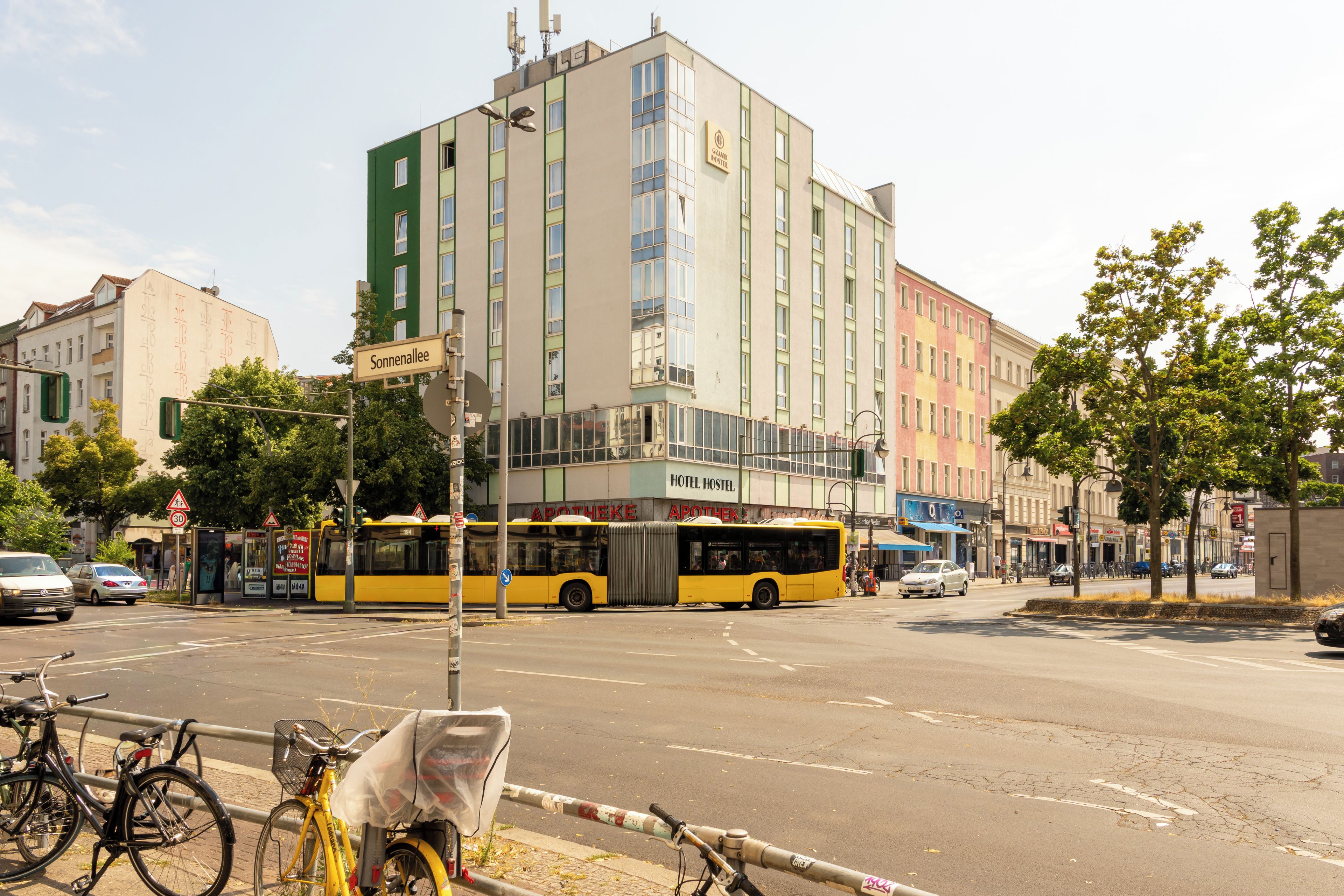 Straßenansicht mit einem gelben Bus, einem Hotel namens 'Grand Hostel' und einem Gebäude mit der Aufschrift 'Sonnenallee'.