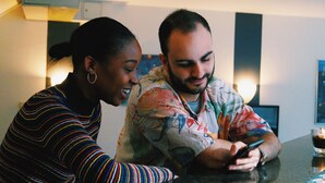Two people sitting at a table with a smartphone, in a room with a modern design and a visible air conditioning unit. - Grand Hostel Berlin Urban (Berlin)