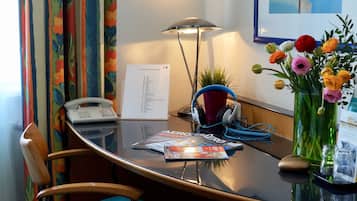Habitación de hotel con escritorio, teléfono, lámpara, auriculares, un florero con flores y un cuadro en la pared.