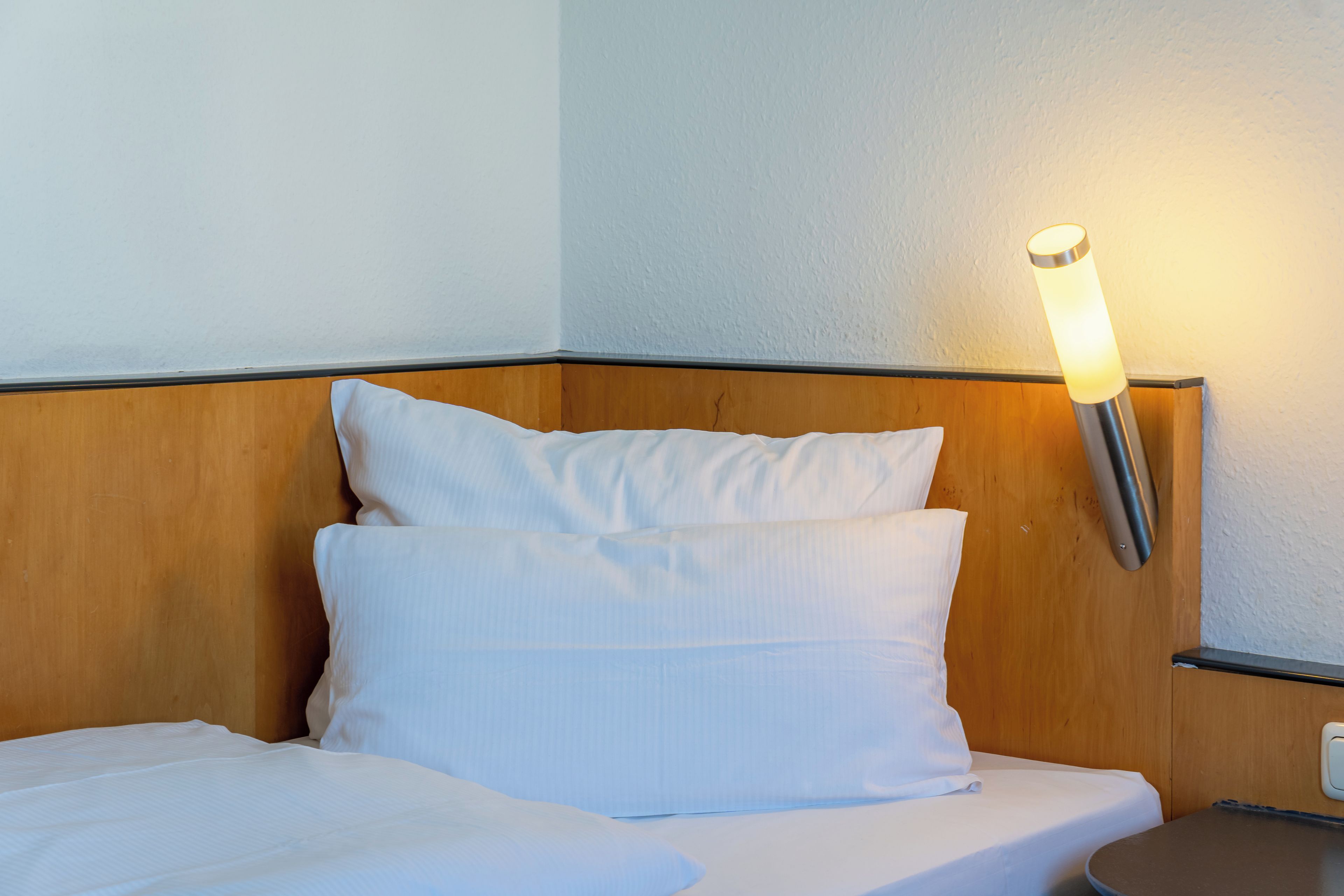 A hotel room with a wooden headboard, a single bed with white bedding, and a wall-mounted light fixture.
