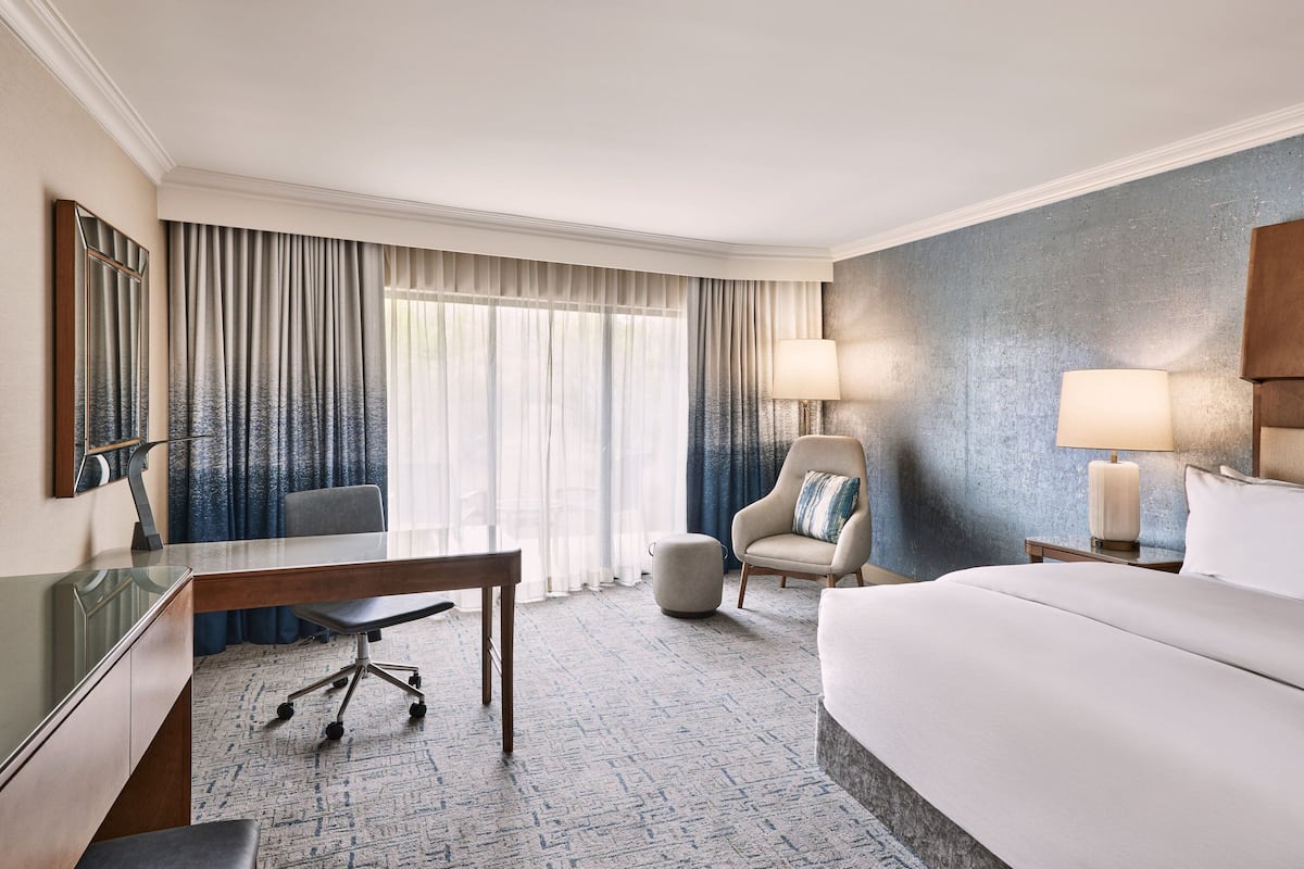 Une chambre d’hôtel avec un grand lit, un bureau avec une chaise, un petit pouf et deux lampes de chevet.