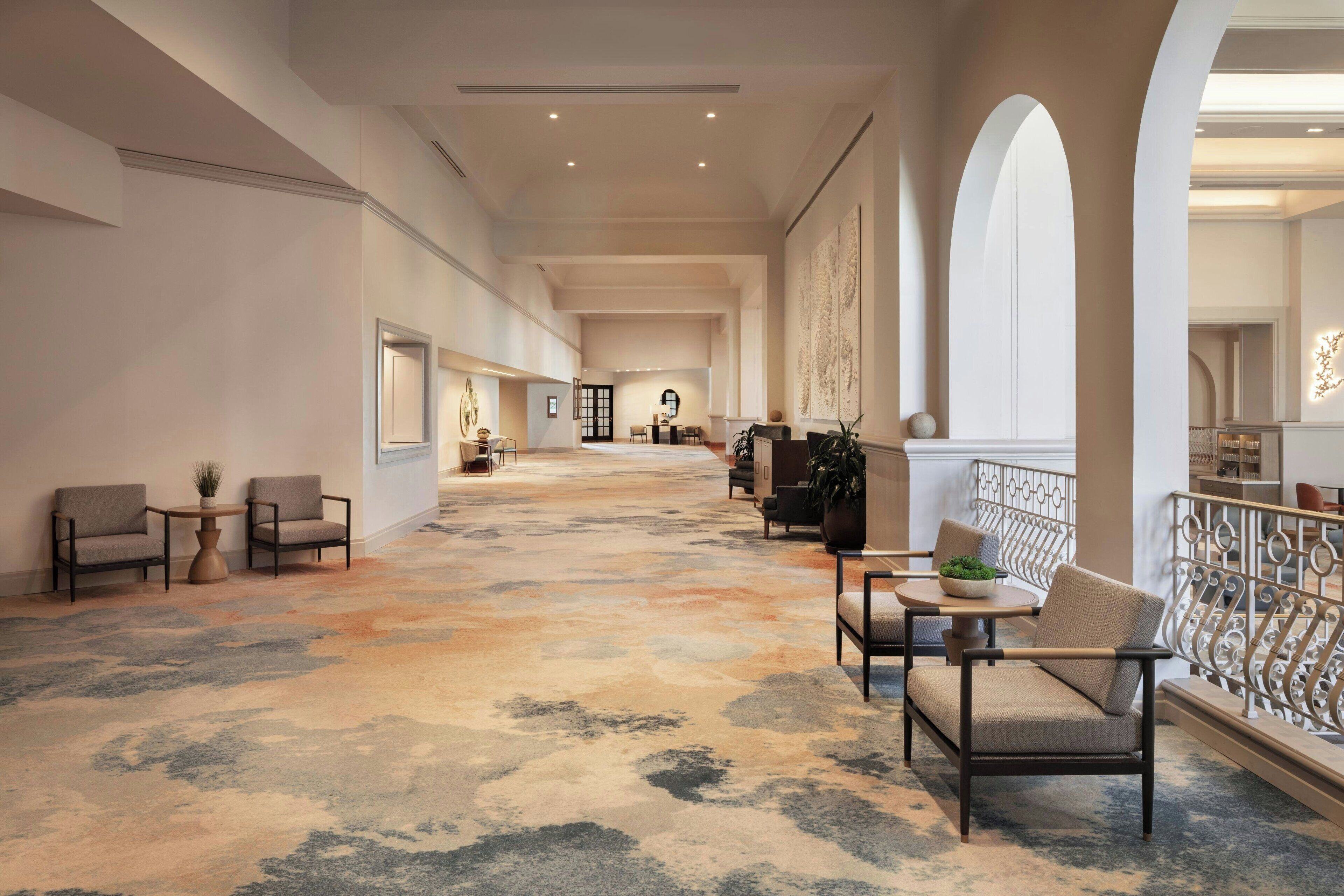 A spacious hallway with a patterned carpet, seating areas with chairs and tables, and a balcony with a railing.