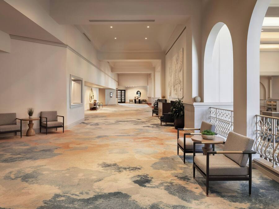 A spacious hallway with a patterned carpet, seating areas with chairs and tables, and a balcony with a railing.