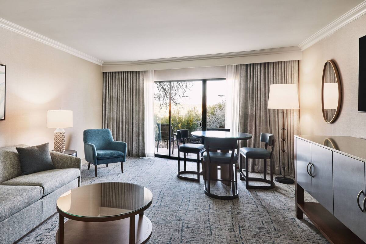 Une chambre d’hôtel avec un canapé, deux fauteuils, une table ronde, un meuble et une grande fenêtre avec des rideaux.
