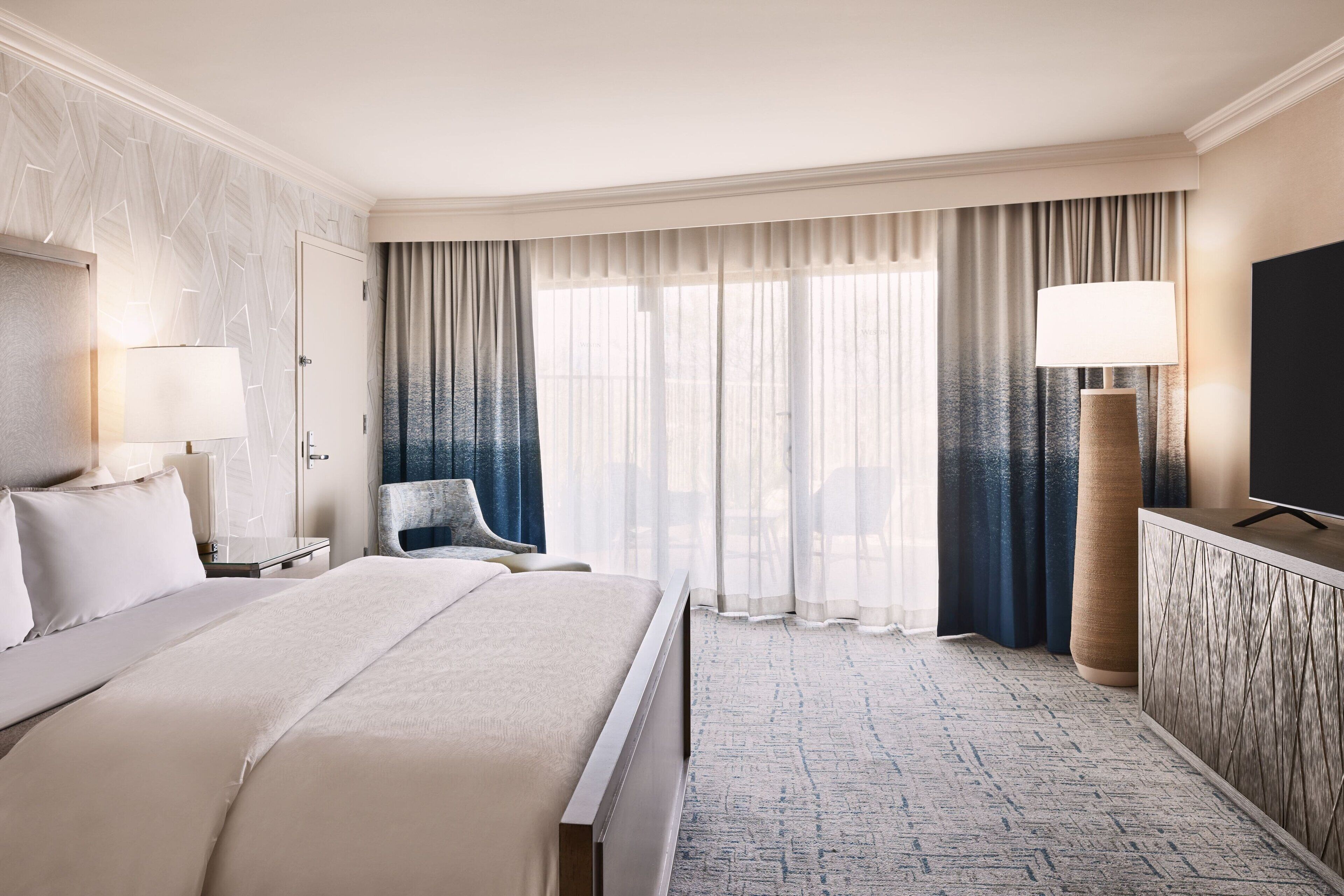 Una habitación de hotel con una cama grande, un escritorio, una silla, un televisor y un ventanal con cortinas.