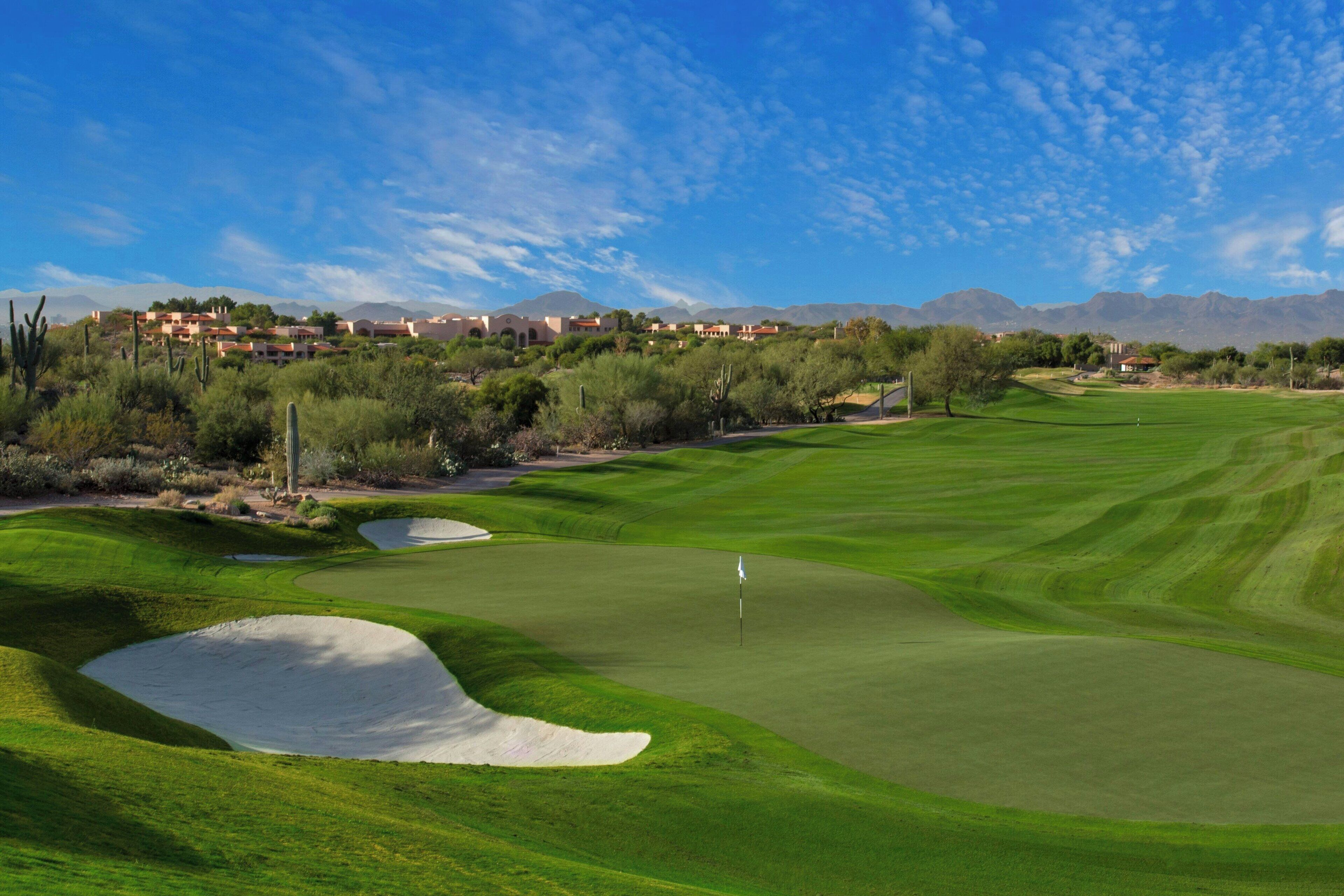 A golf course with bunkers, a flagstick, and distant buildings.