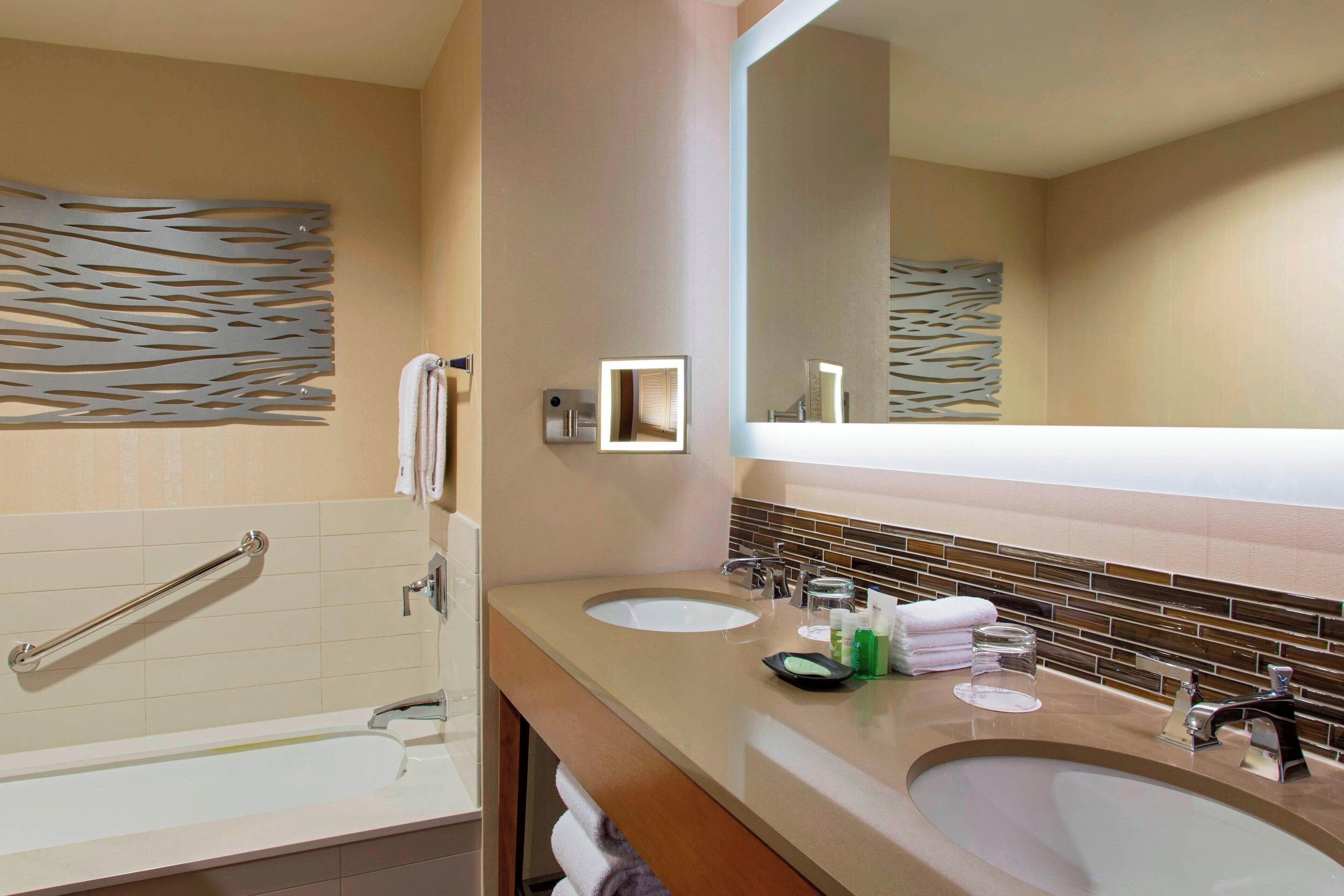 A modern bathroom with two sinks, a large mirror, and a grab bar in the shower area.