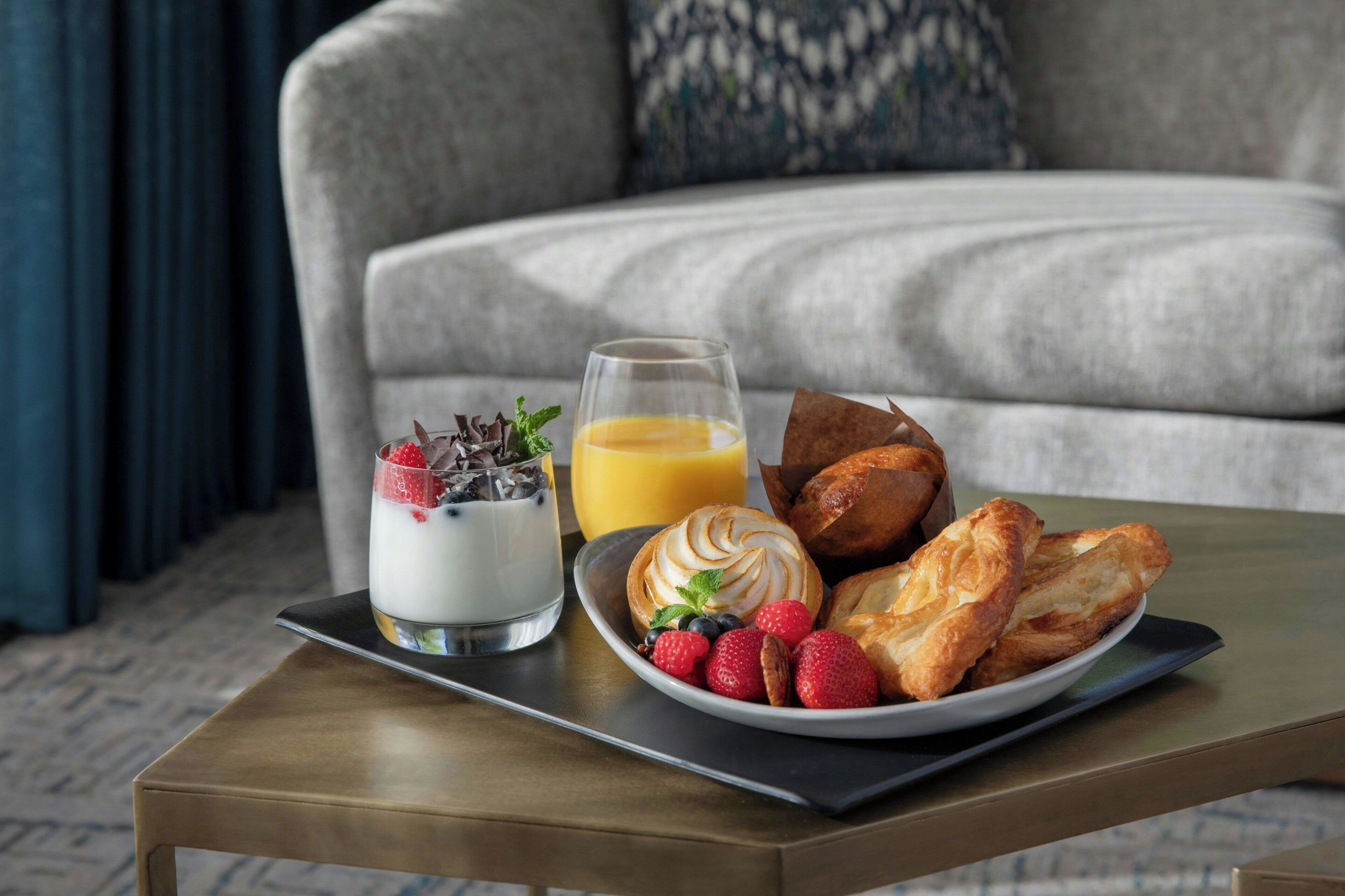 A breakfast tray with yogurt, orange juice, pastries, and berries on a table in a living room with a sofa and curtains.