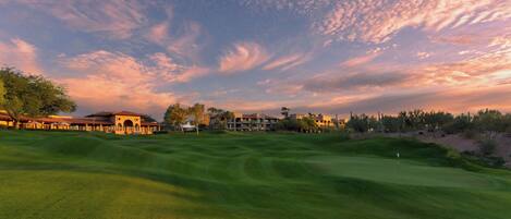 A golf course with a clubhouse, a putting green, and a backdrop of trees and buildings.