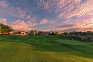 A golf course with a clubhouse, a putting green, and a backdrop of trees and buildings.