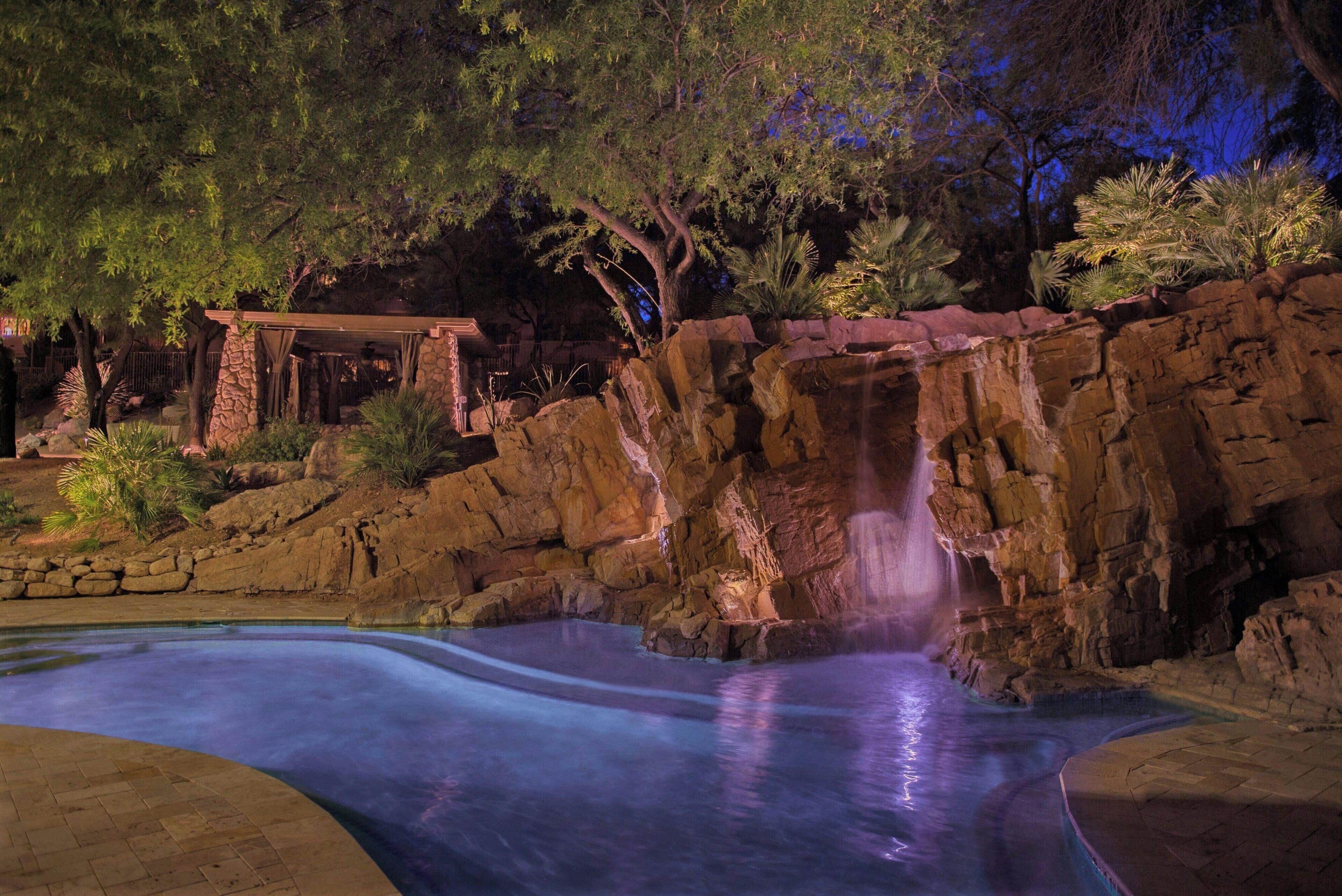 Zona de piscina exterior de nit amb cascada, envoltada de roques i arbres.