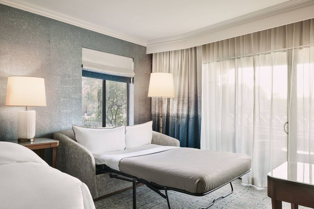 Une chambre d’hôtel avec un lit, deux tables de chevet, une lampe, une fenêtre avec des rideaux et une vue sur la verdure.