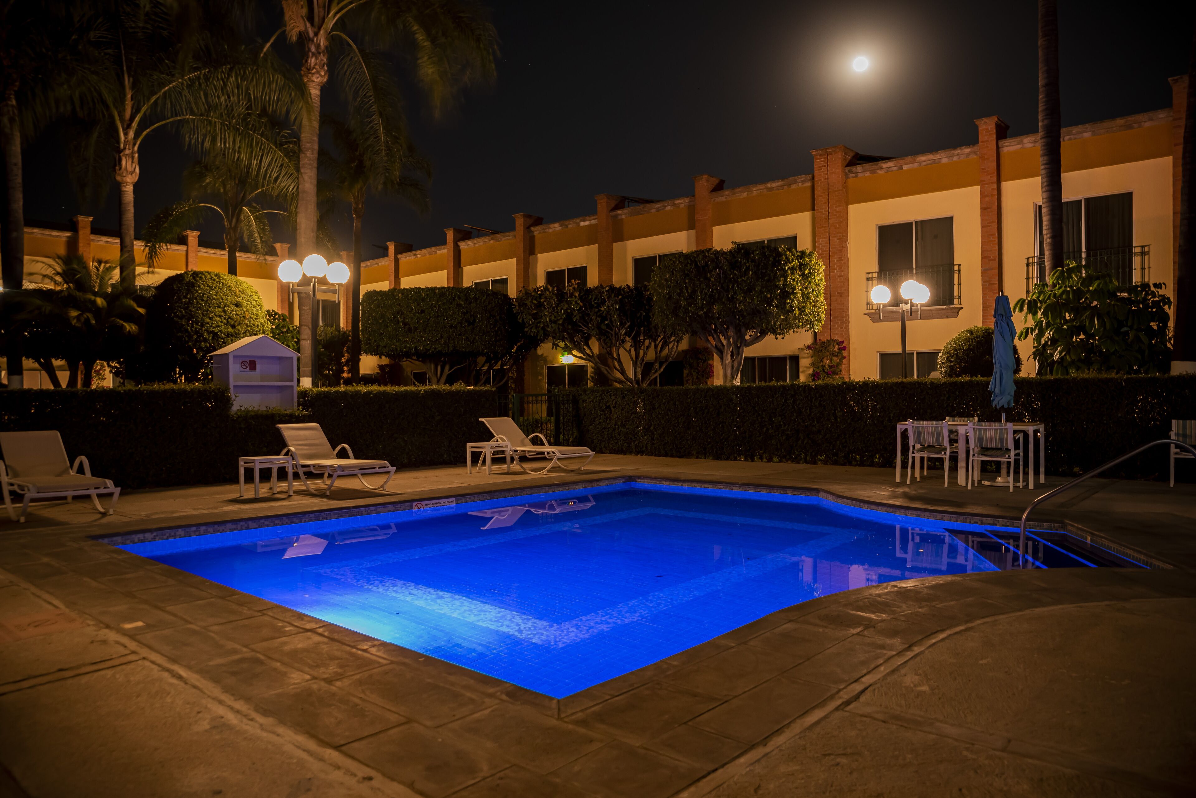 A swimming pool with lounge chairs and a small table, surrounded by palm trees and a building with balconies.