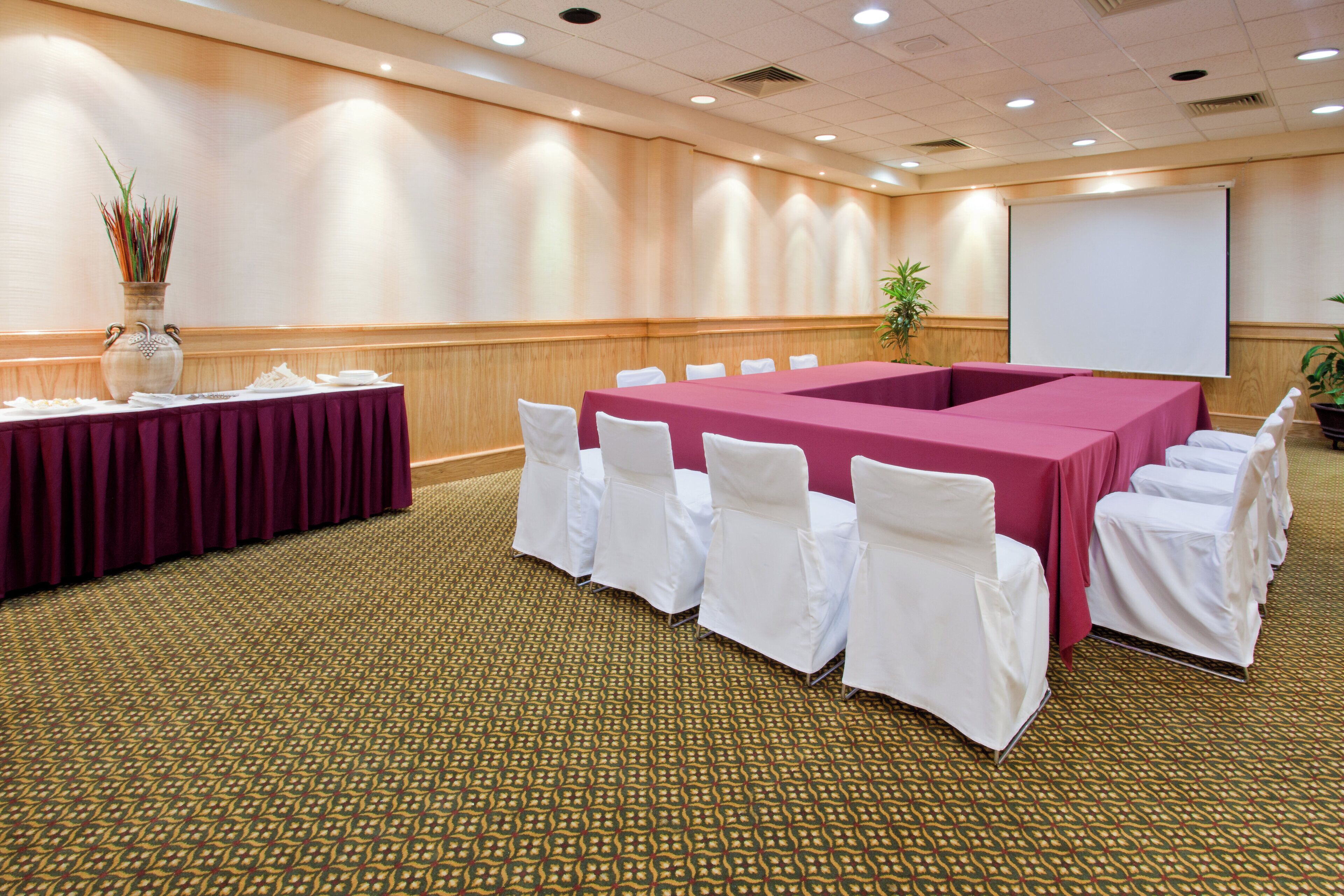 Een vergaderzaal met een lange tafel, witte stoelen, een scherm voor een beamer en een plant.