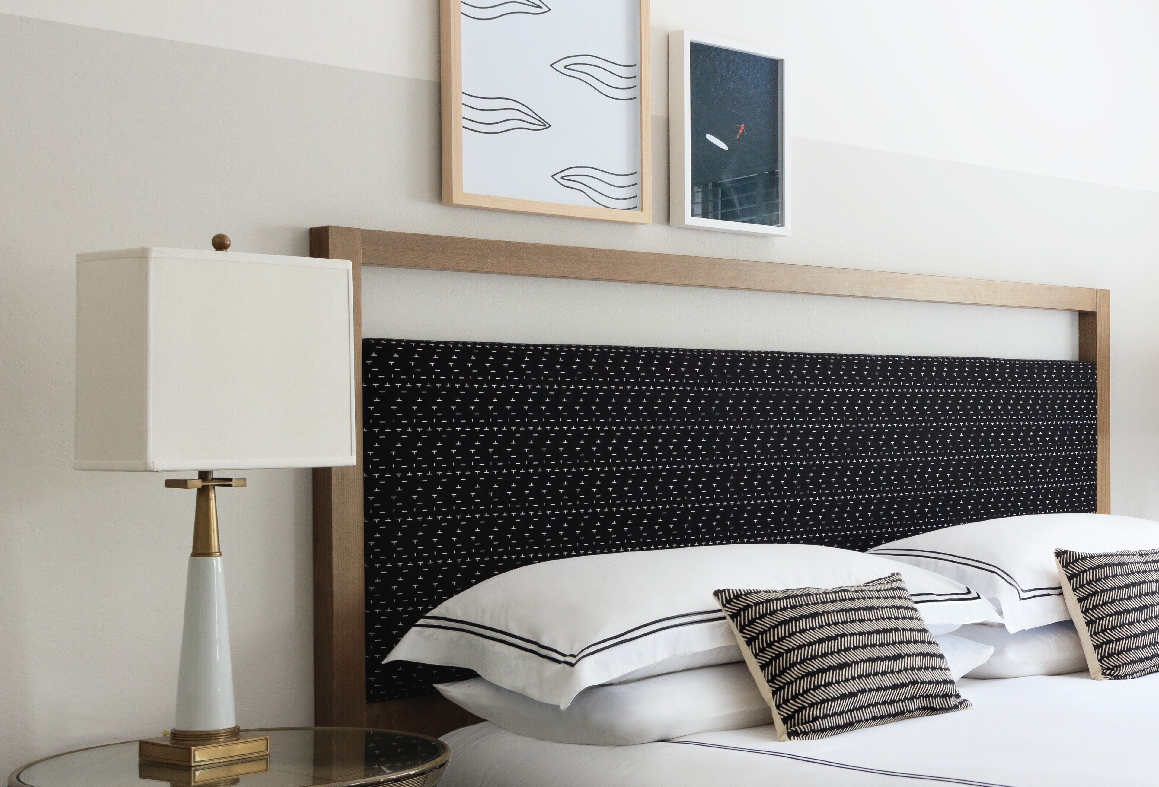 Una cama bien hecha con almohadas blancas y negras, un cabecero con estampado blanco y negro, y una lámpara de noche sobre una mesita de vidrio.