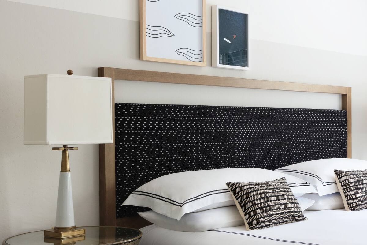 Una cama bien hecha con almohadas blancas y negras, un cabecero con estampado blanco y negro, y una lámpara de noche sobre una mesita de vidrio.