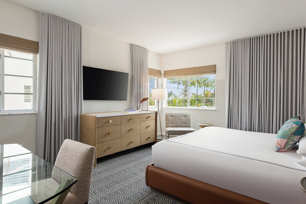 A hotel room with a bed, a desk, a chair, a dresser, a TV, and a window with a view.