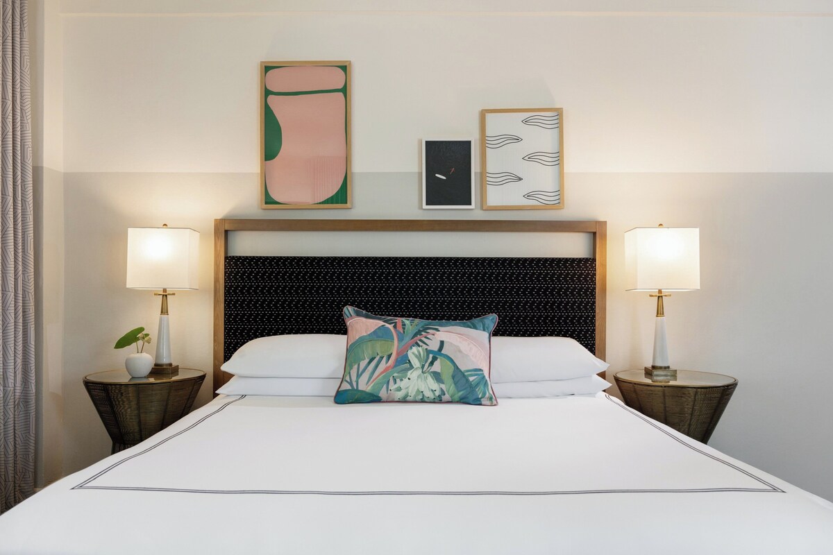 A hotel room with a large bed, two bedside tables with lamps, a headboard with a textured pattern, and wall art above the bed.
