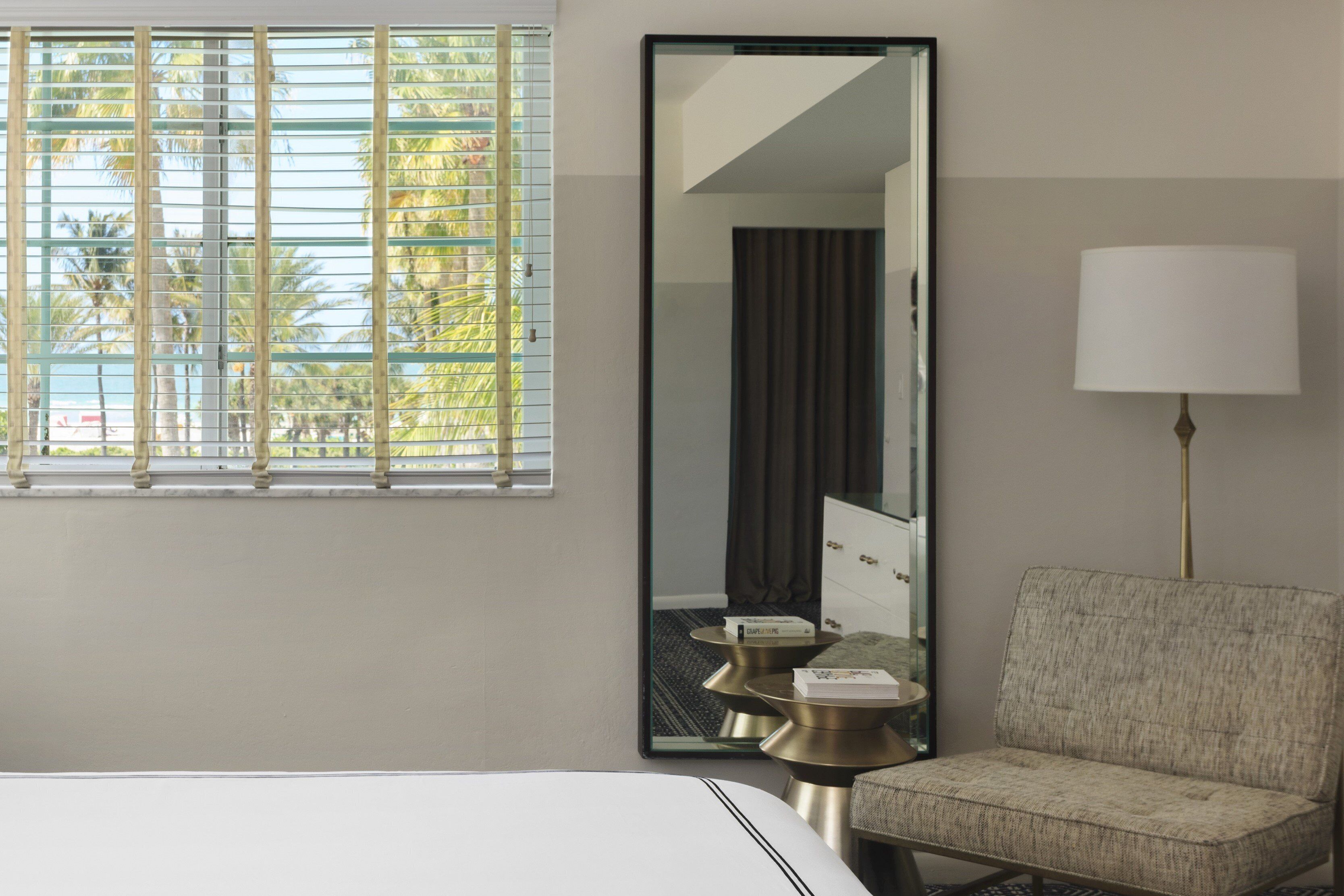 Habitación de hotel con cama, una silla, lámpara, espejo y ventana con persianas.