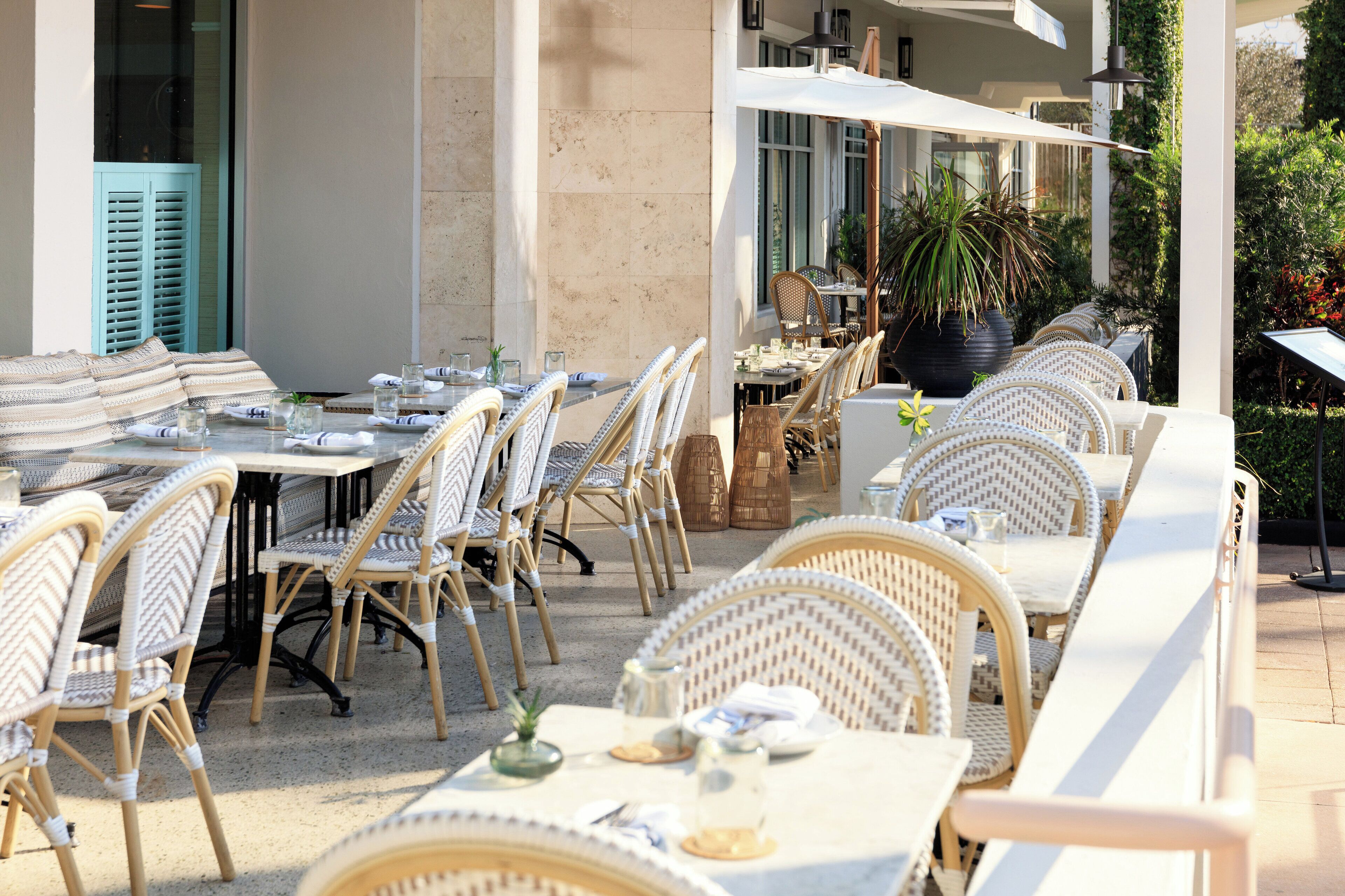 Un espace extérieur aménagé pour les repas, avec des chaises et des tables en osier, entouré d’un bâtiment doté de grandes fenêtres et agrémenté d’une plante en pot.