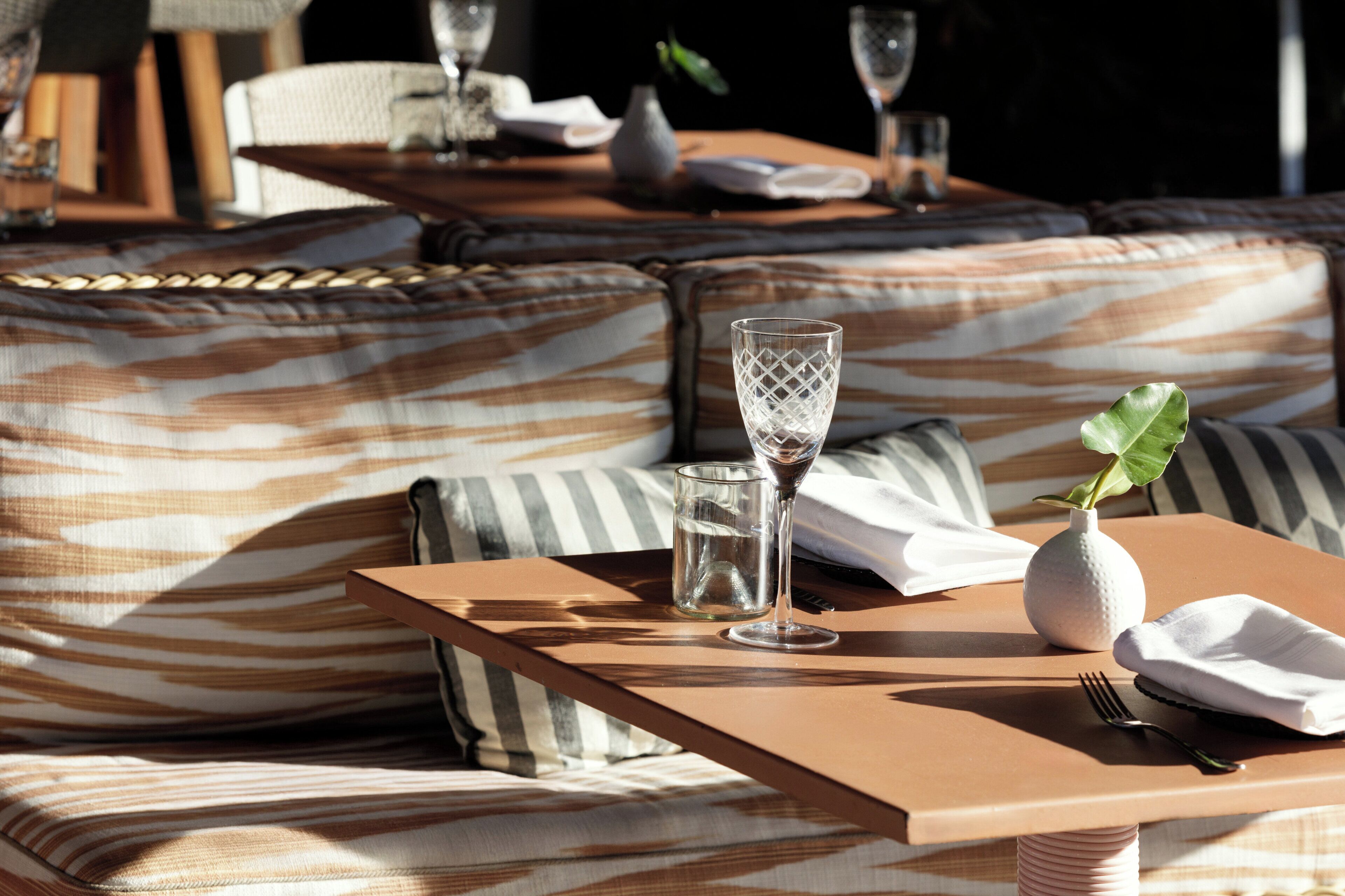 Een zonnig buitenterrein met een tafel, kussens, een glas en een vaas.