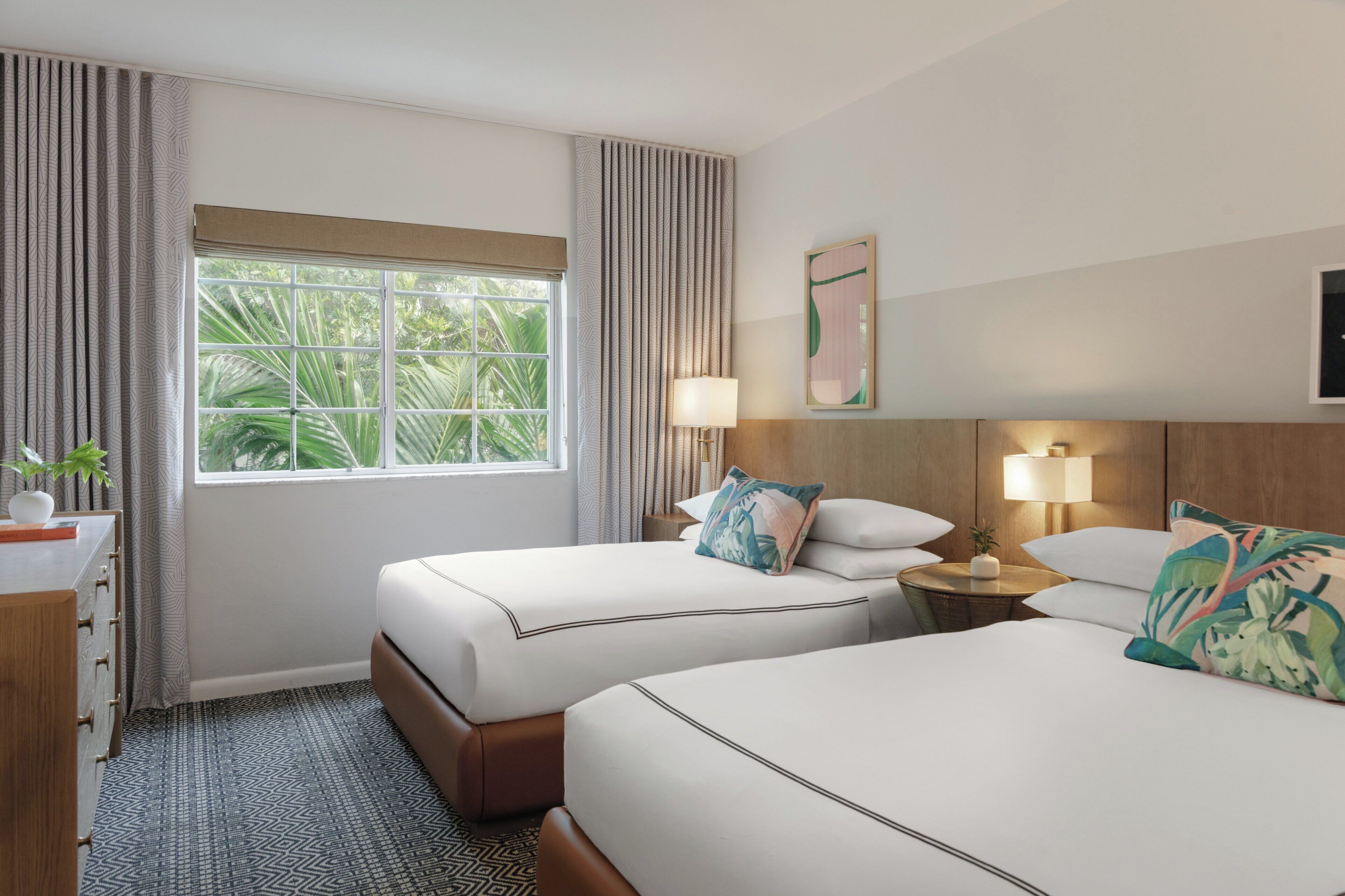 Habitación de hotel con dos camas, un escritorio, una lámpara y vistas al exterior con vegetación.