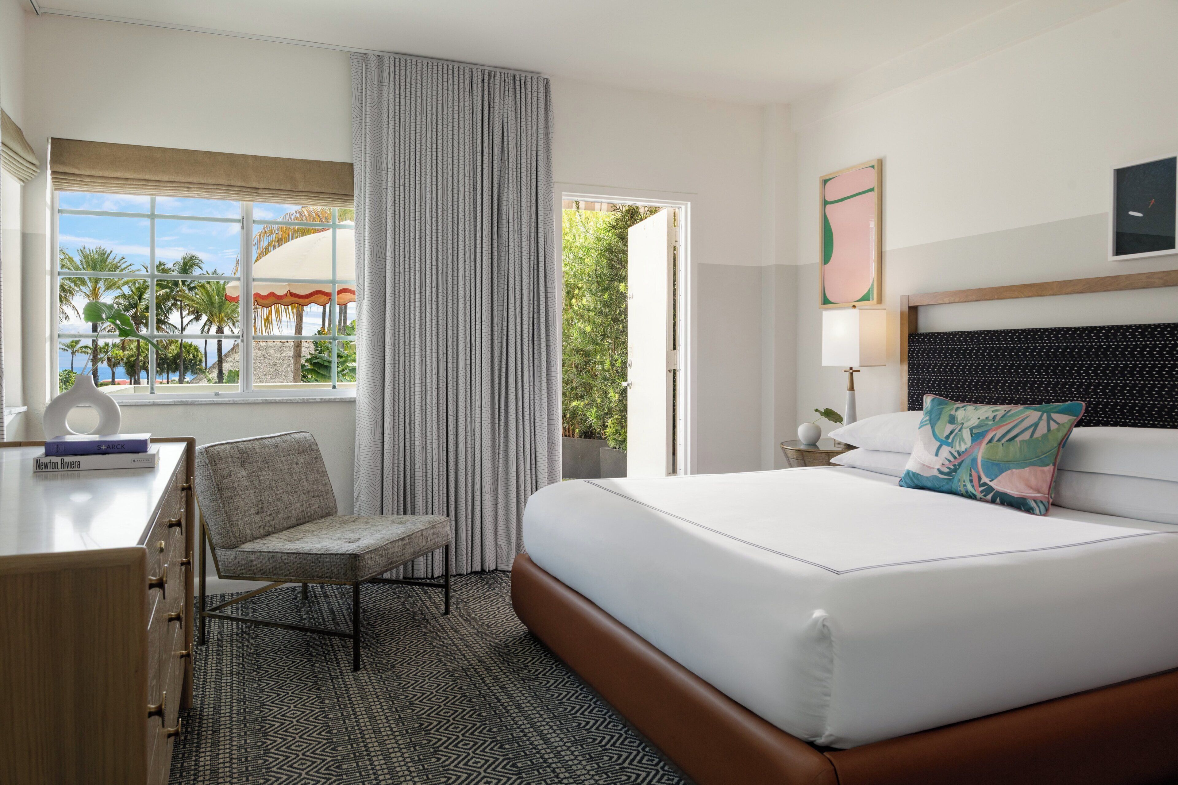 Une chambre d’hôtel comprenant un lit, un bureau, une chaise, une fenêtre donnant sur des palmiers et une plage, ainsi qu’une porte menant à l’extérieur.
