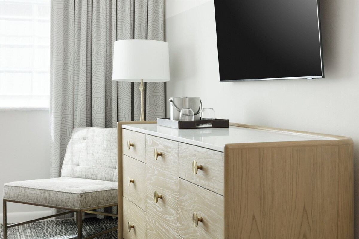 A modern hotel room with a wooden dresser, a flat-screen TV, a lamp, and a chair.