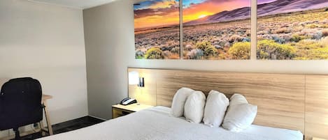 Quarto de hotel com uma cama grande, cabeceira de madeira, escrivaninha com cadeira e mesa de cabeceira.