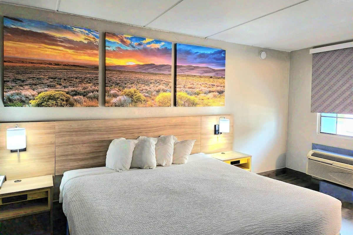 A hotel room with a large bed, two bedside tables, a nightstand, a wall-mounted lamp, and a large landscape painting above the bed.