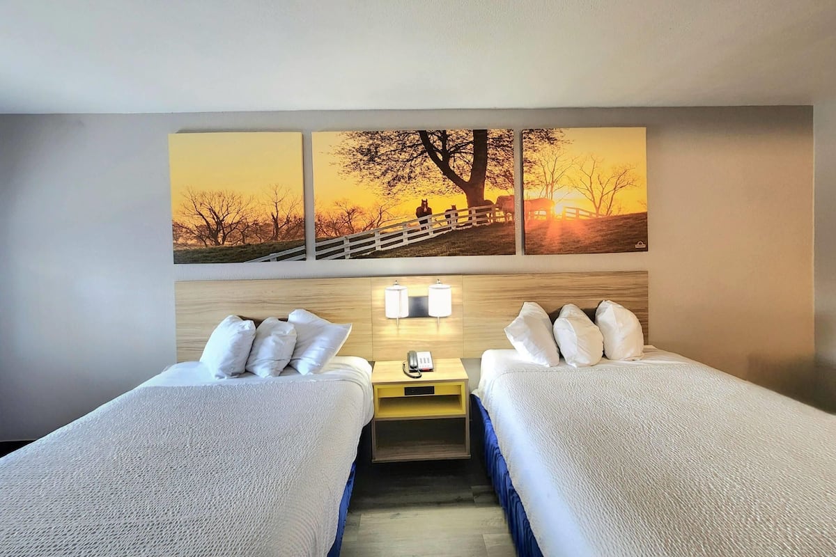 A hotel room with two beds, a wooden headboard, a nightstand with a phone, and a sunset painting above the beds.