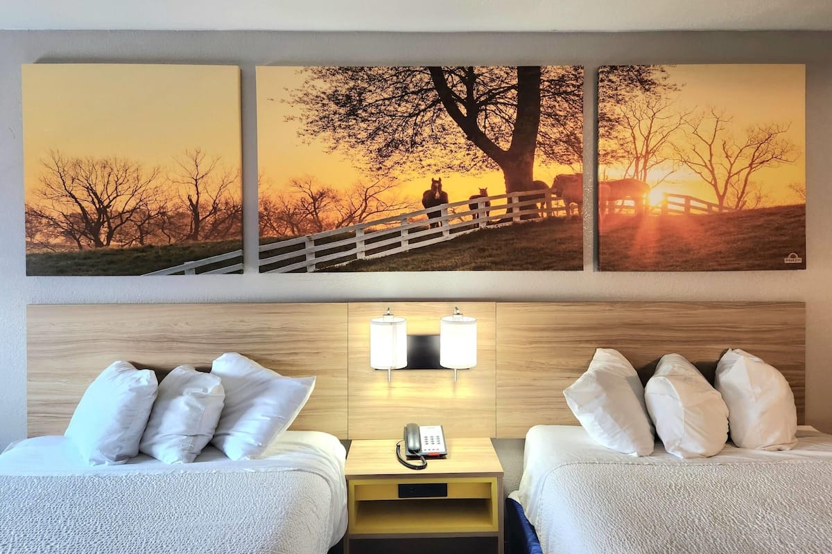 A hotel room with two beds, a wooden headboard, a nightstand with a phone, and a wall-mounted lamp.
