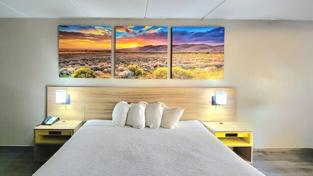 A hotel room with a large bed, two bedside tables, a nightstand with a phone, and a wall-mounted painting of a sunset landscape. - Days Inn by Wyndham Houston (Houston)
