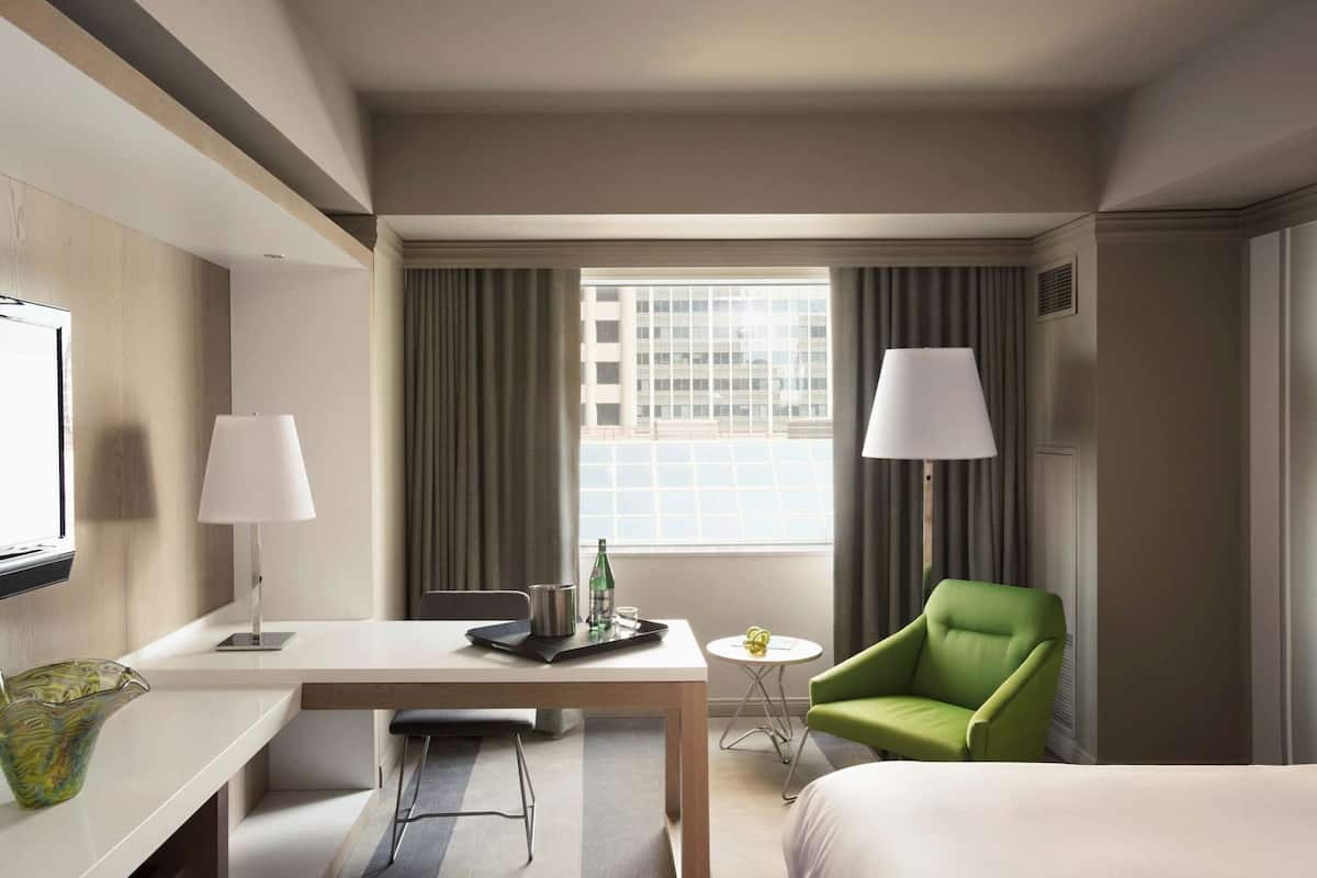 A hotel room with a bed, a desk with a chair, a small table with a lamp, and a green armchair.
